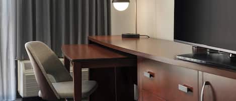A hotel room with a wooden desk, a chair, a flat-screen TV, a desk lamp, and a built-in heater.