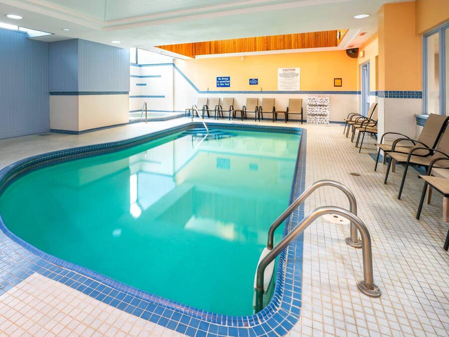An indoor swimming pool with clear blue water, tiled floor, and a poolside area with chairs and a wooden ceiling.