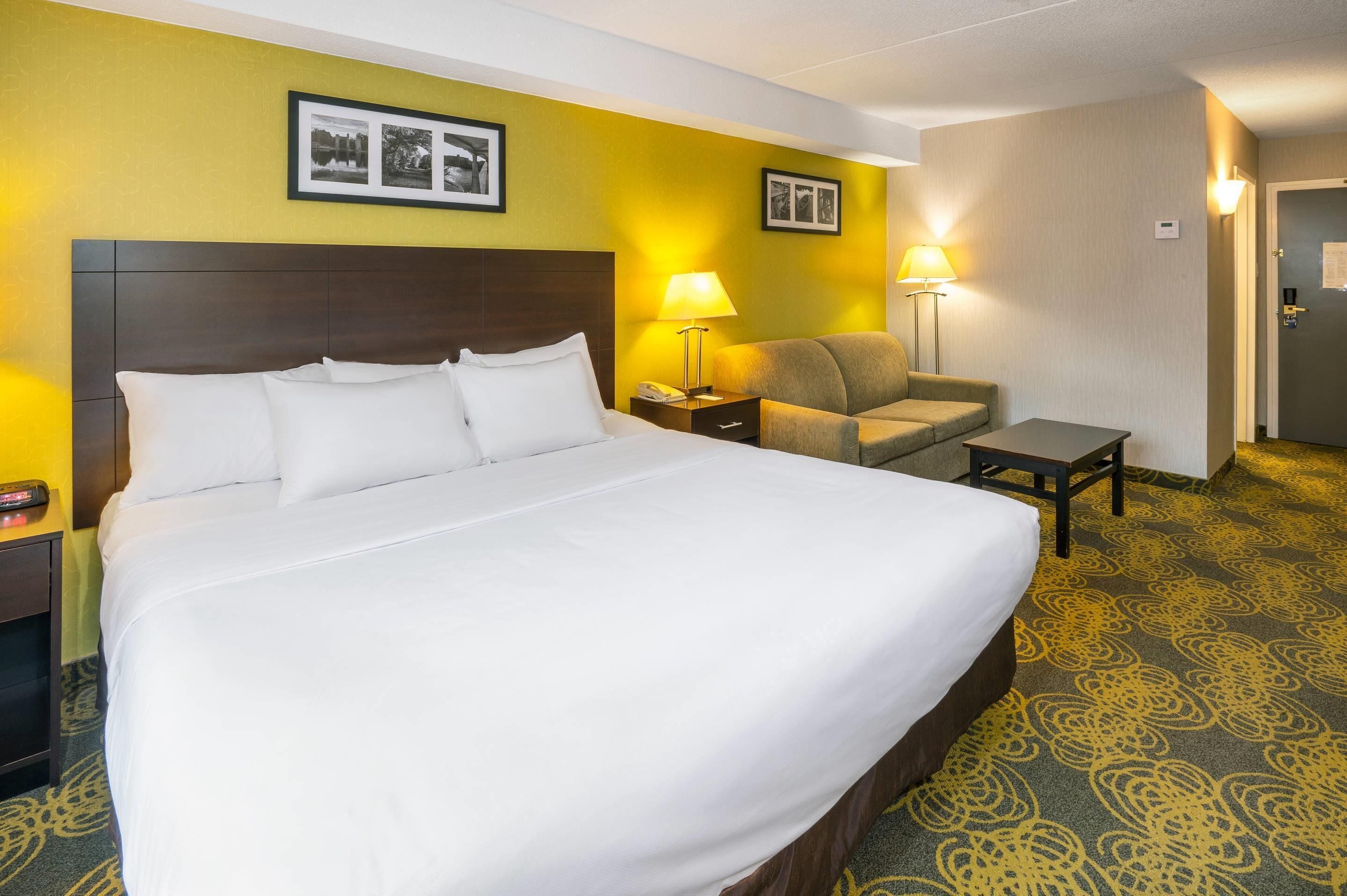 A hotel room with a large bed, a bedside table with a lamp, a chair, and a small table.