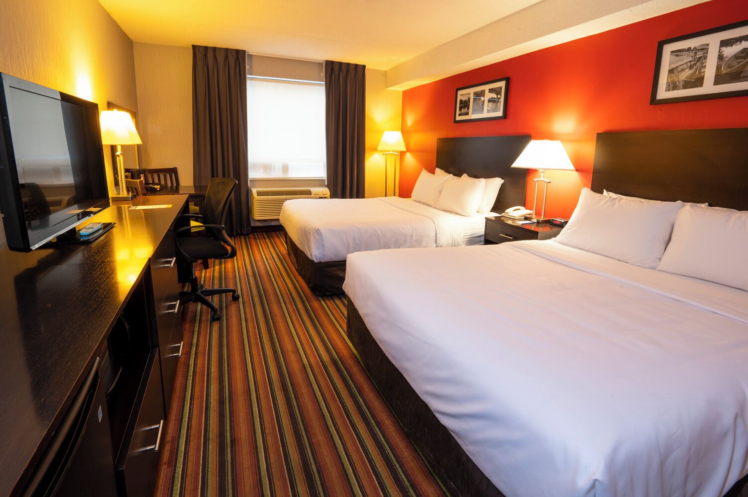 A hotel room with two beds, a desk with a TV, a desk lamp, a chair, and a window with curtains.