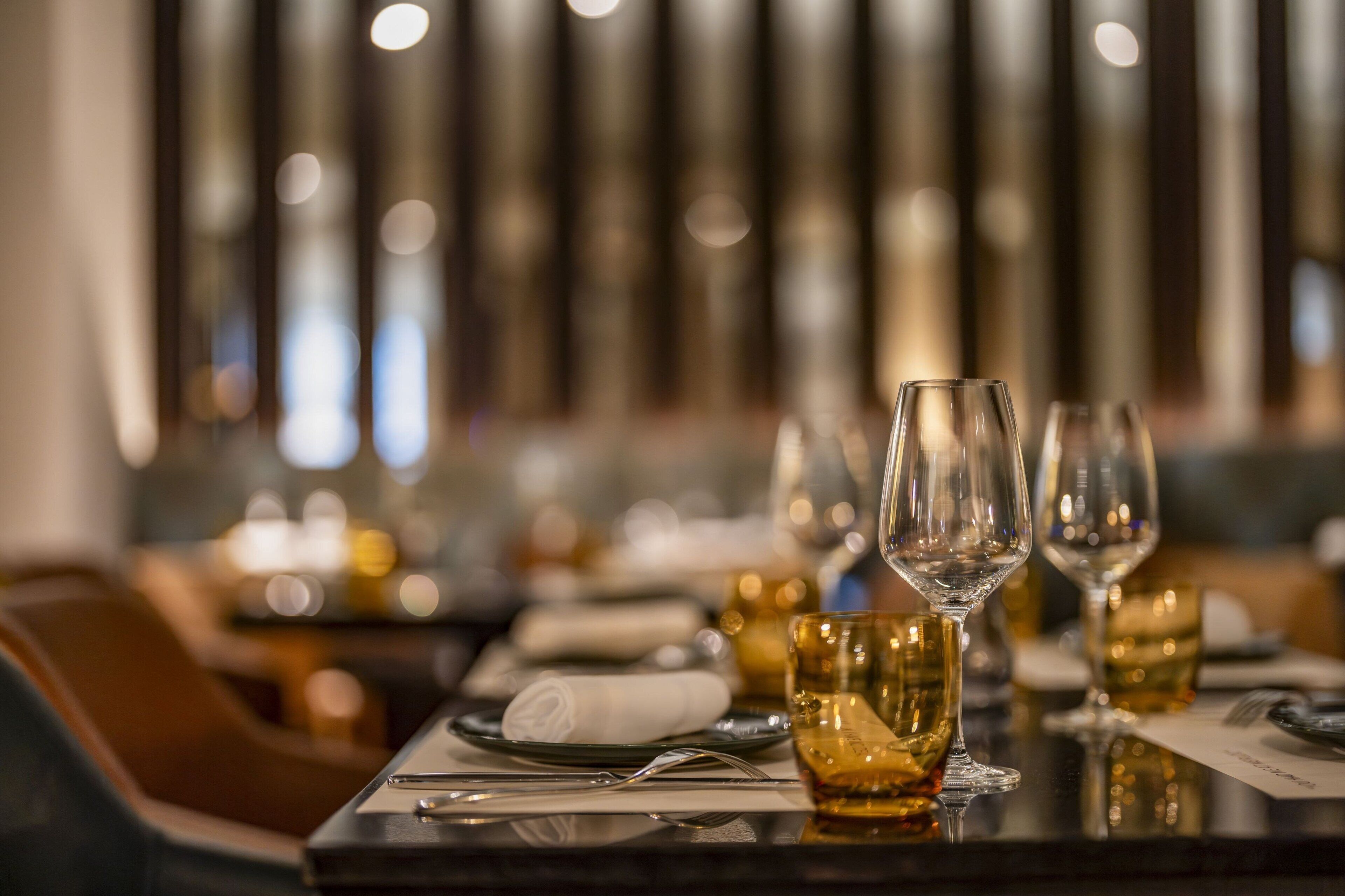 A restaurant setting with a table set for dining, featuring wine glasses and a glass of amber-colored beverage.