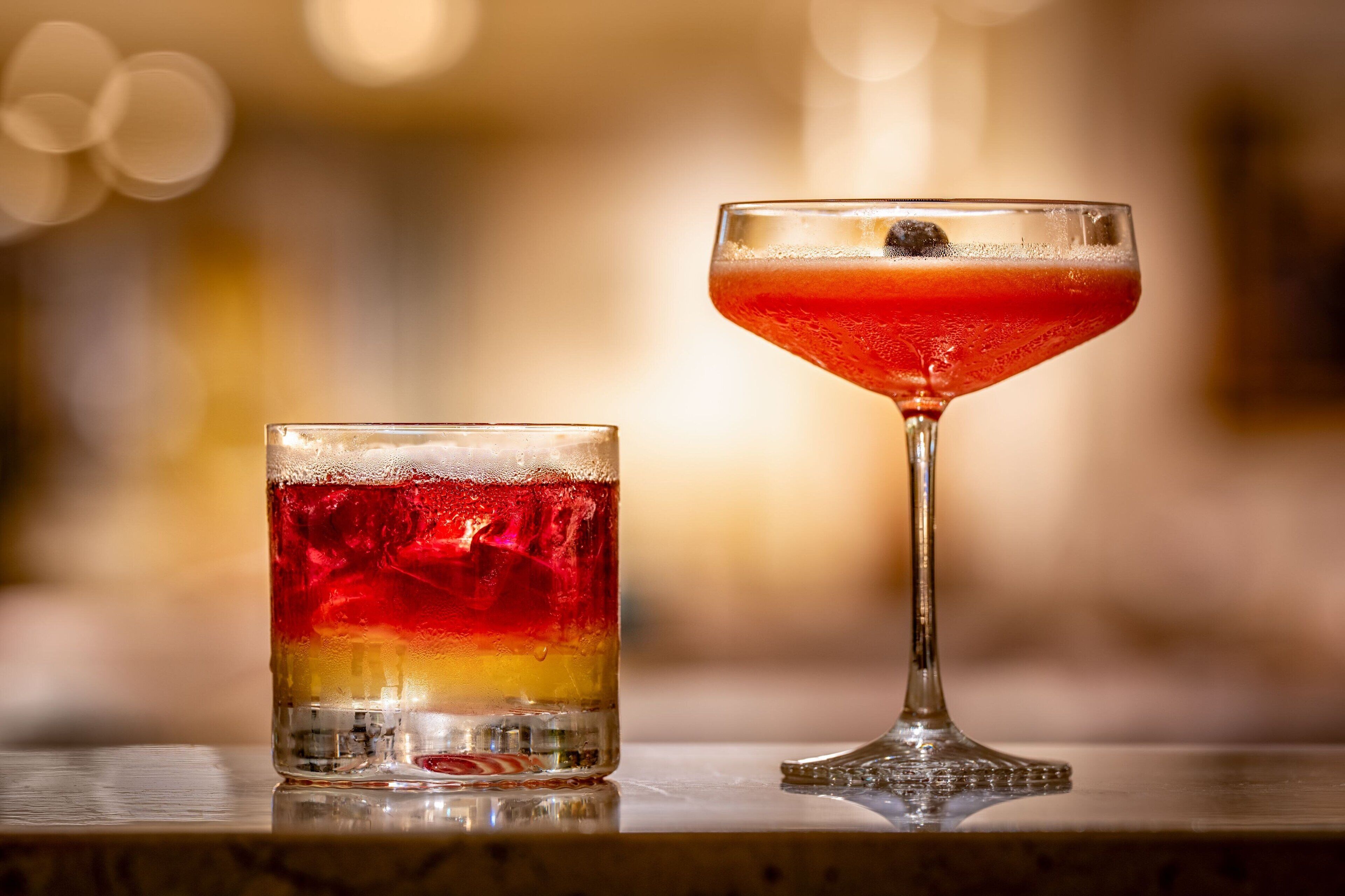 Two glasses of a layered beverage, one in a short glass and one in a stemmed glass, on a reflective surface.