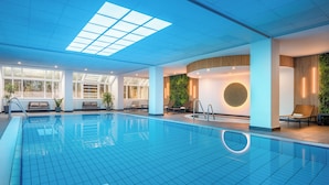 An indoor swimming pool with clear blue tiles, a skylight ceiling, and modern seating areas.
