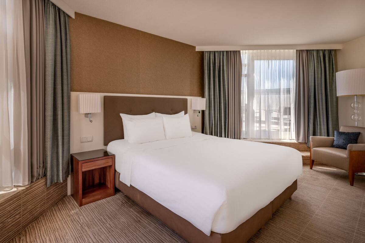 A hotel room with a large bed, a nightstand, a chair, and a window with curtains.