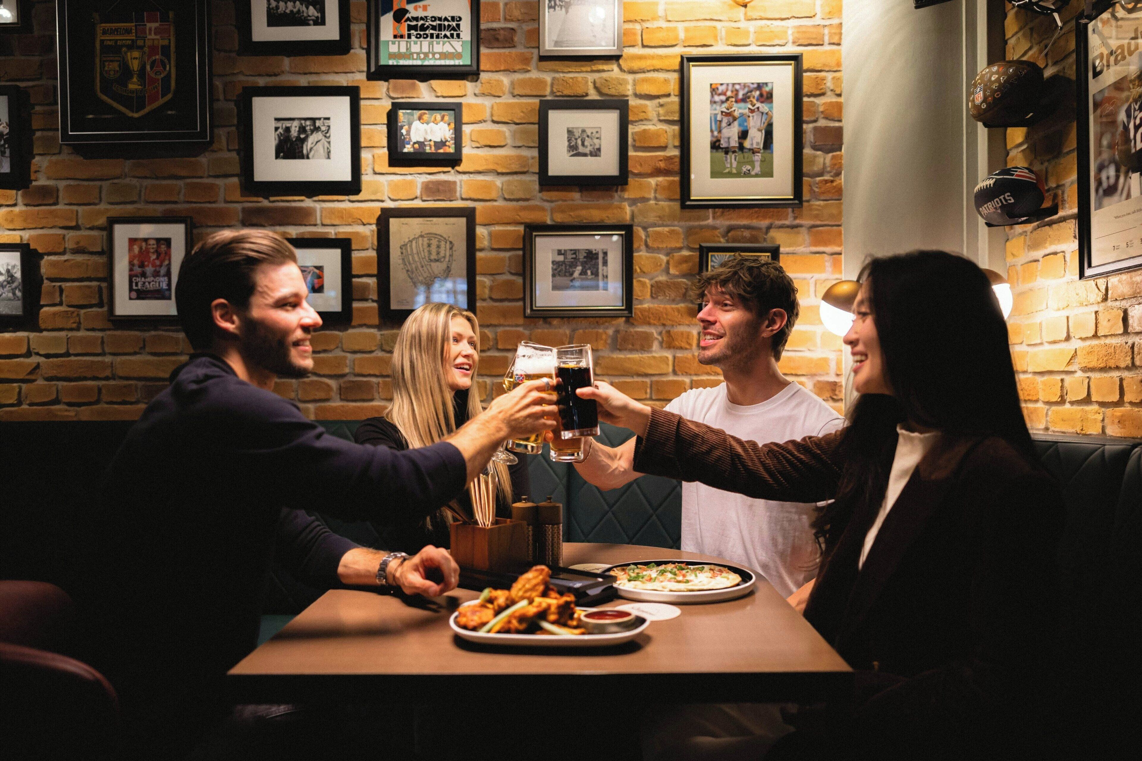 A group of people toasting at a table with food and drinks in a brick-walled room with framed pictures on the wall.