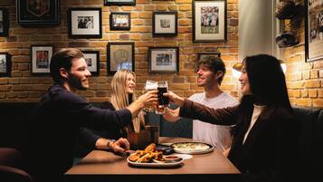 A group of people toasting at a table with food and drinks in a brick-walled room with framed pictures on the wall.