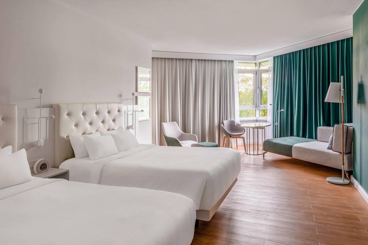 A hotel room with two beds, a sitting area with a sofa and chairs, a small table, and a window with curtains.