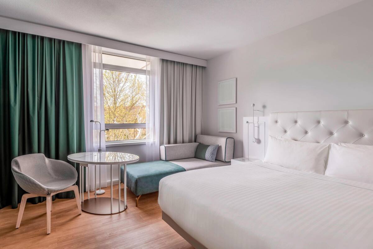 A modern hotel room with a large bed, a seating area with a table, and a view of trees through the window.