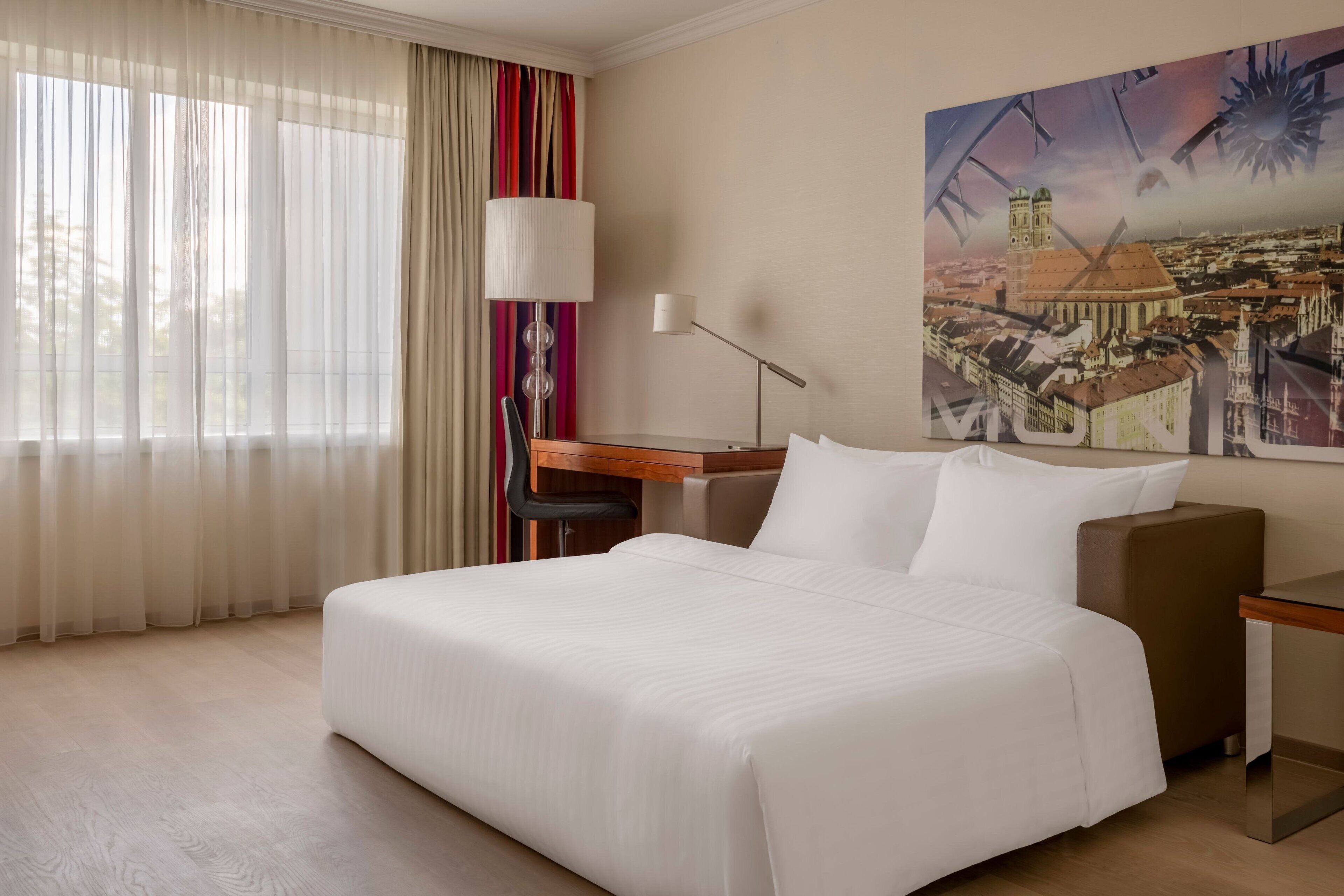 A hotel room with a large bed, a desk, a chair, a lamp, and a painting of a cityscape on the wall.