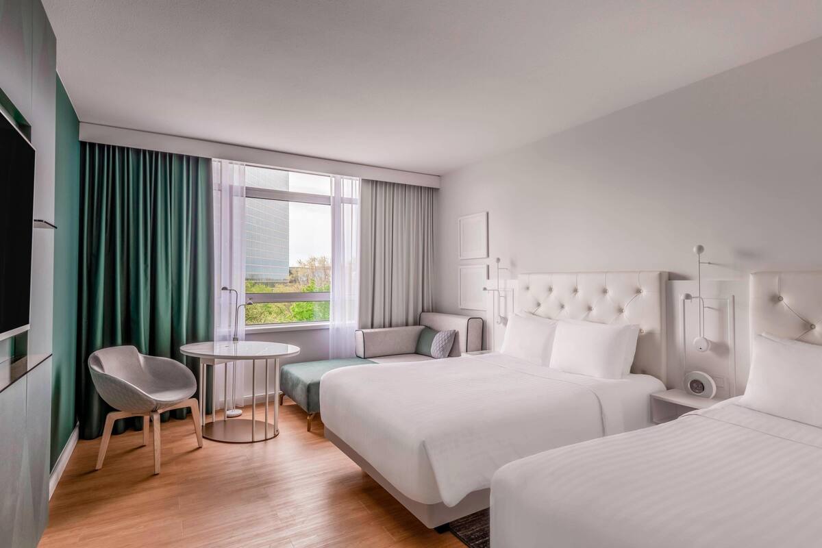 A modern hotel room with two beds, a small round table, a chair, and a view of greenery through large windows.