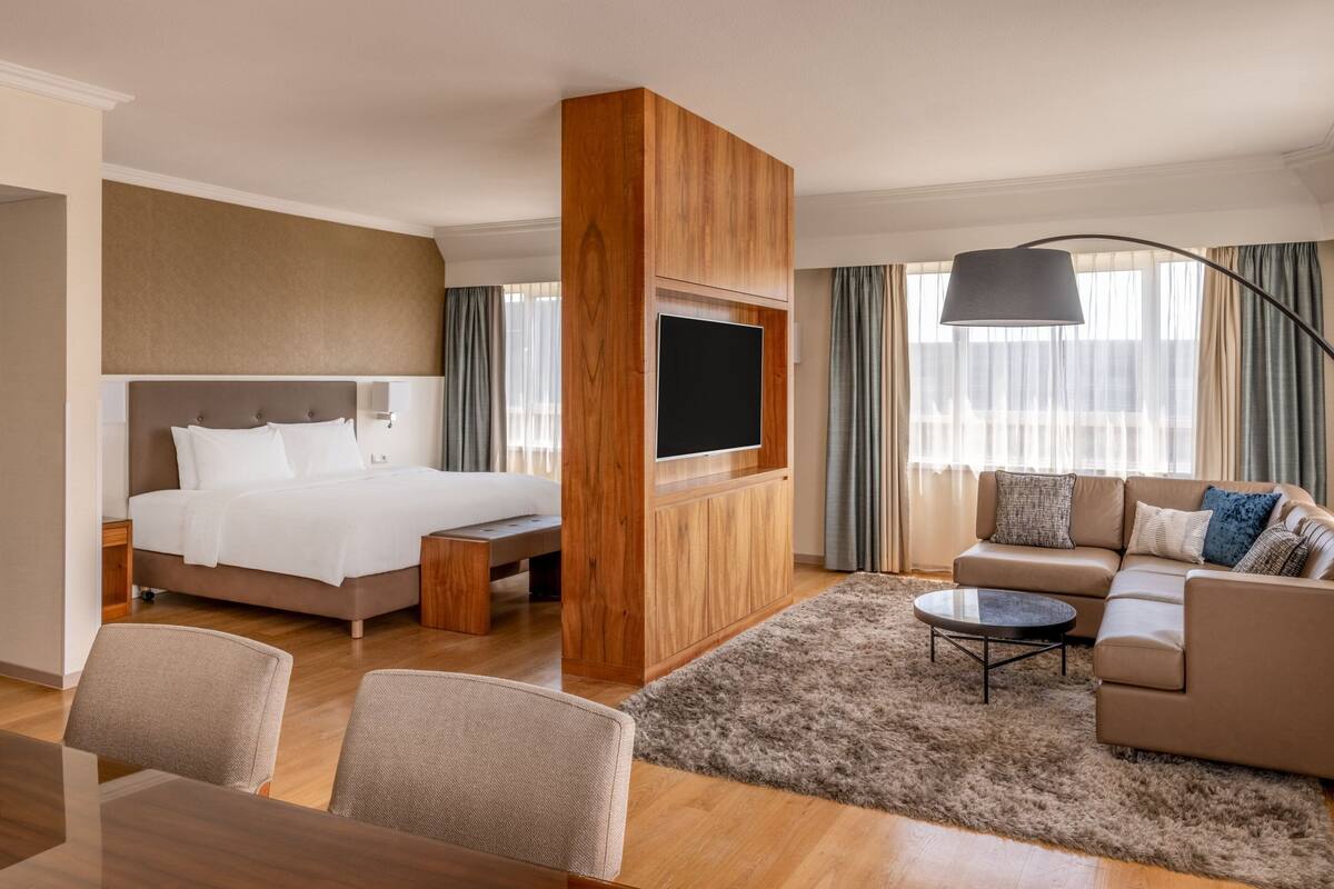 A hotel room with a large bed, a sofa, a TV mounted on a wooden panel, a dining table with chairs, and a large window with curtains.