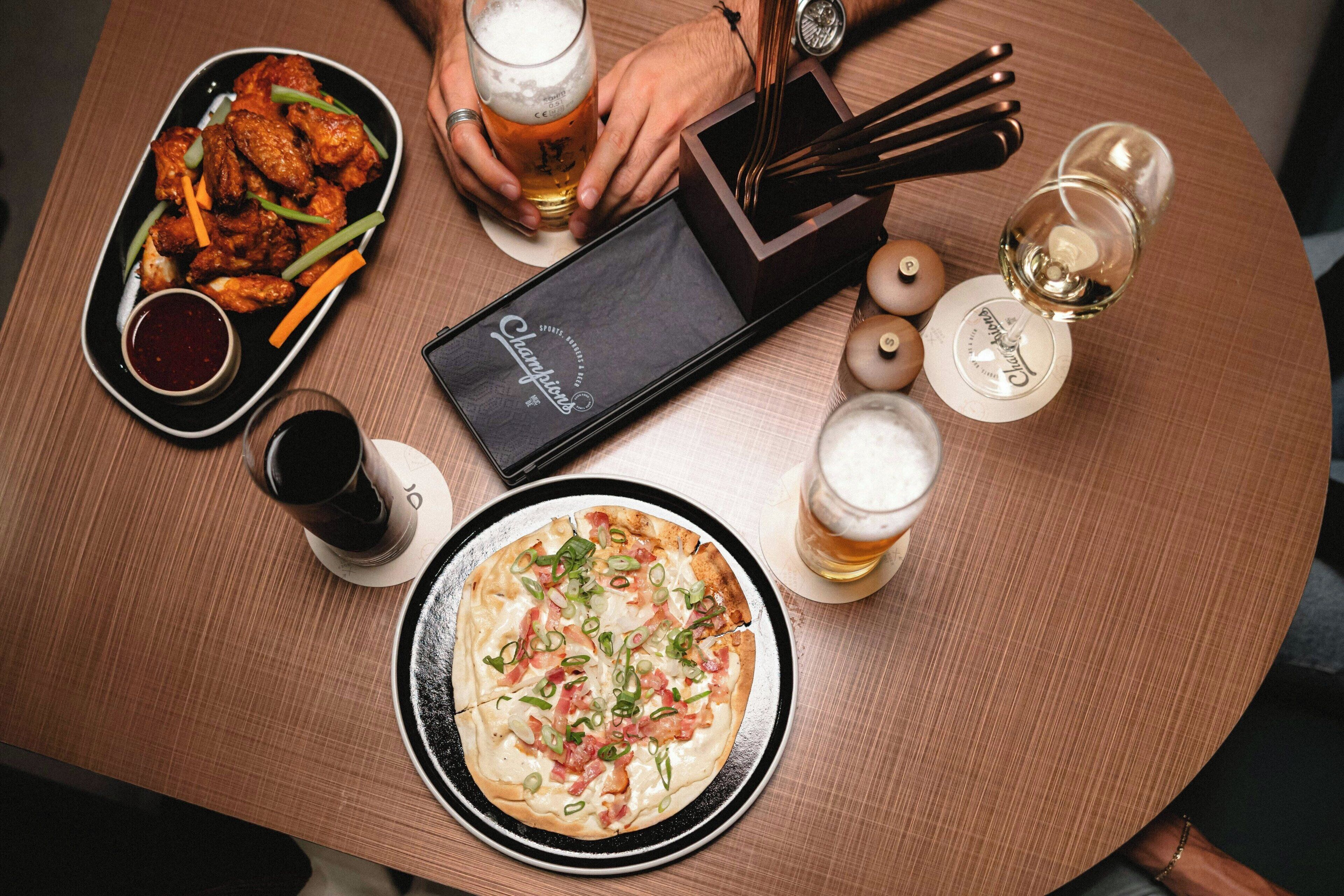 Una mesa con una pizza, una bandeja de alitas de pollo, una cerveza en un vaso y una botella de vino.