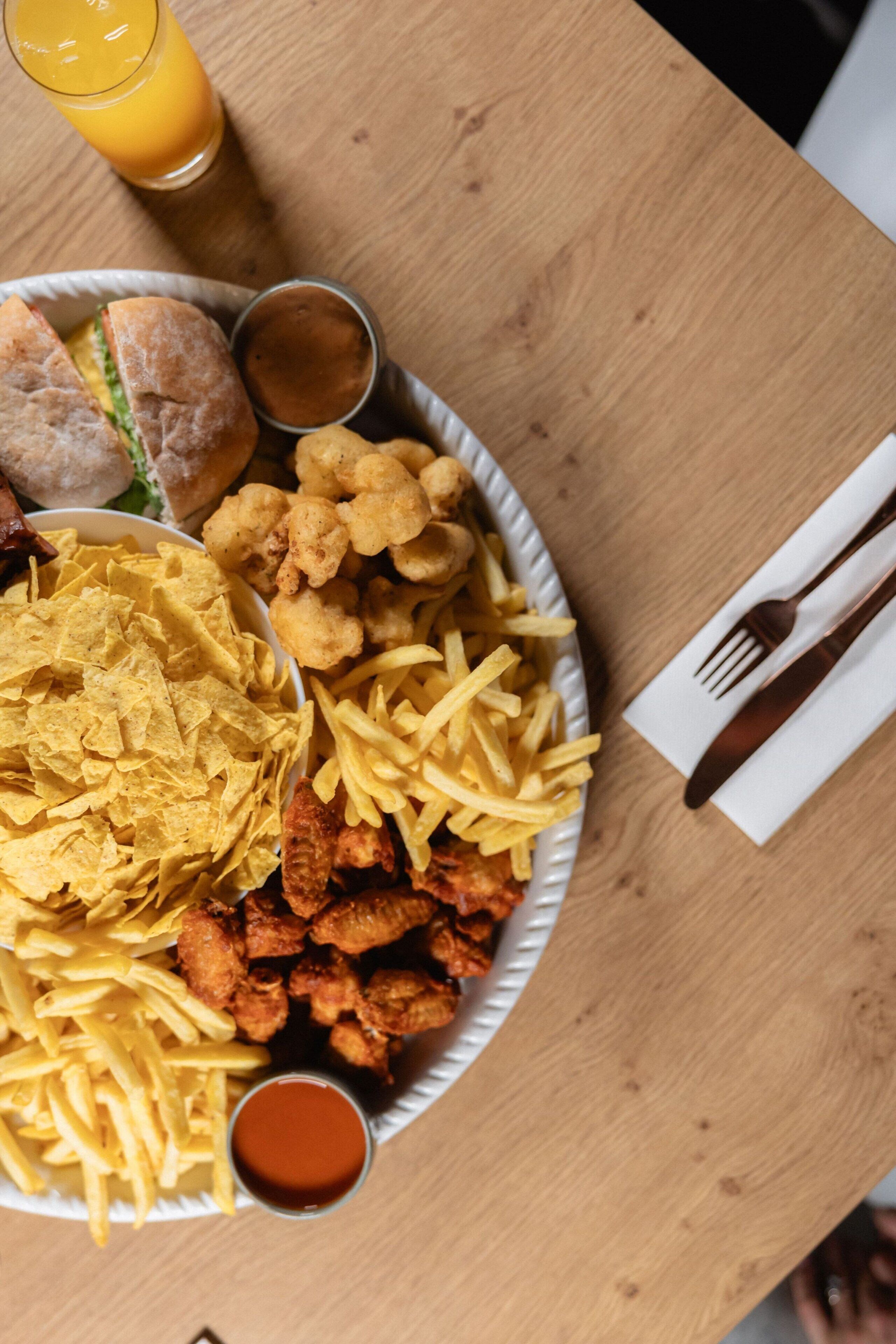 En tallrik med mat som innehåller pommes frites, kycklingvingar och en smörgås, tillsammans med en dryck och en servett på ett träbord.