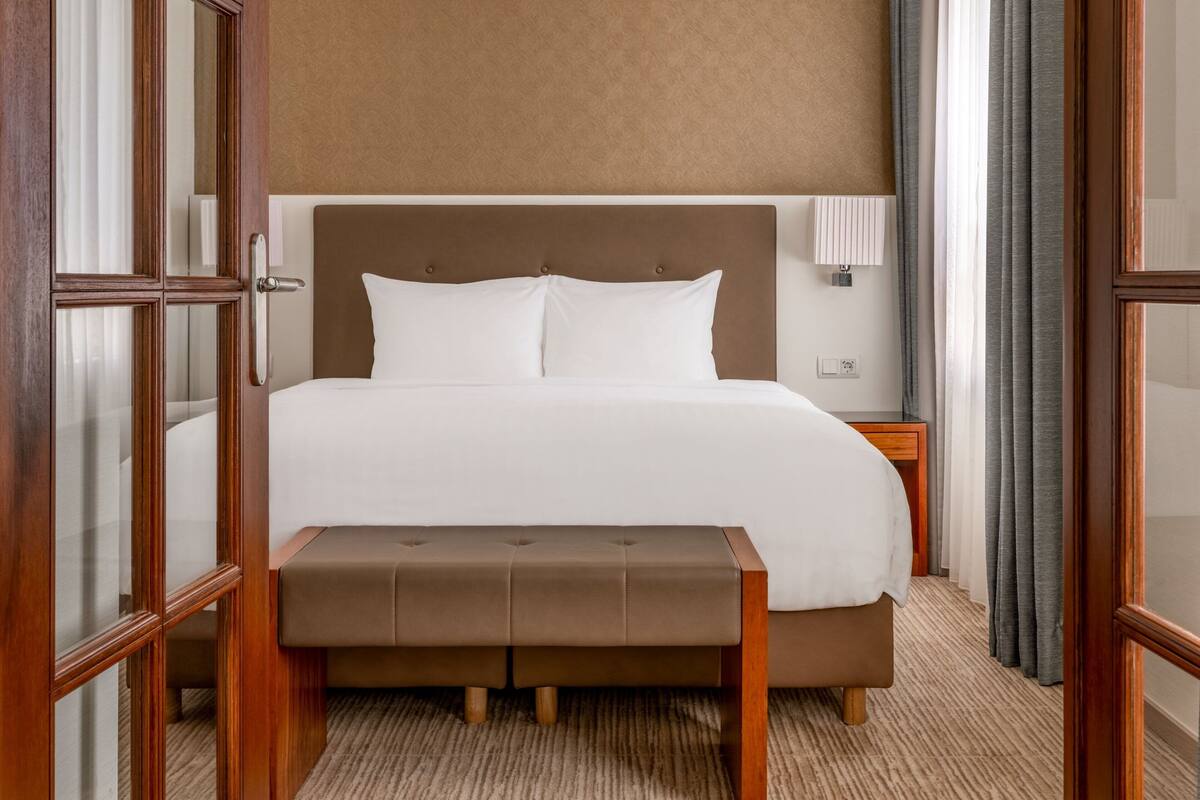 A hotel room with a large bed, a wooden bench, a nightstand, and a wall-mounted lamp.