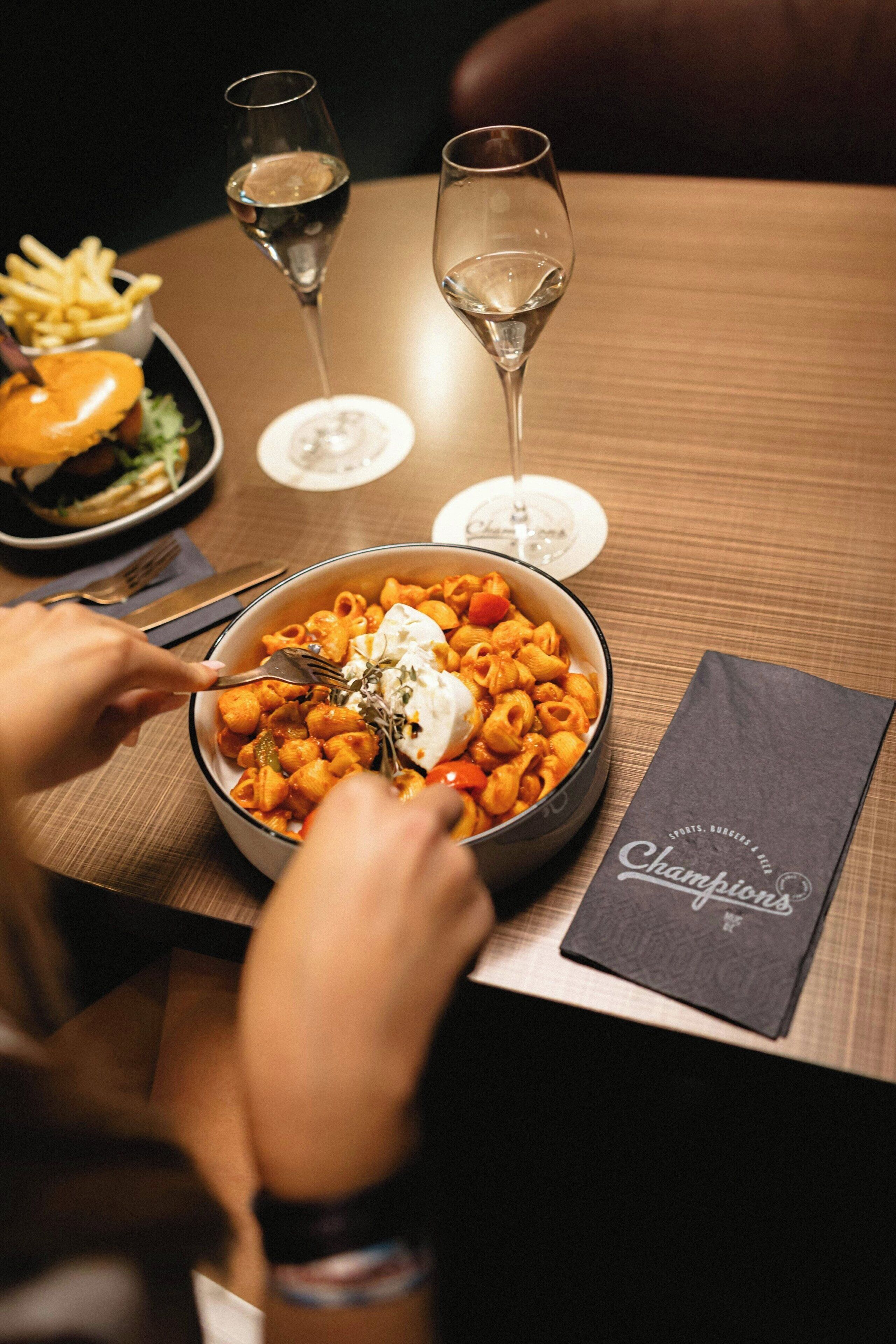 A dining table with a bowl of pasta, a burger, and two glasses of wine.