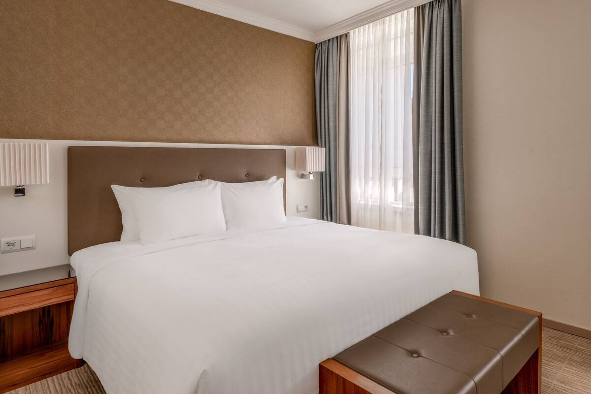 A neatly made bed with white linens, a wooden nightstand with a lamp, and a small wooden bench.