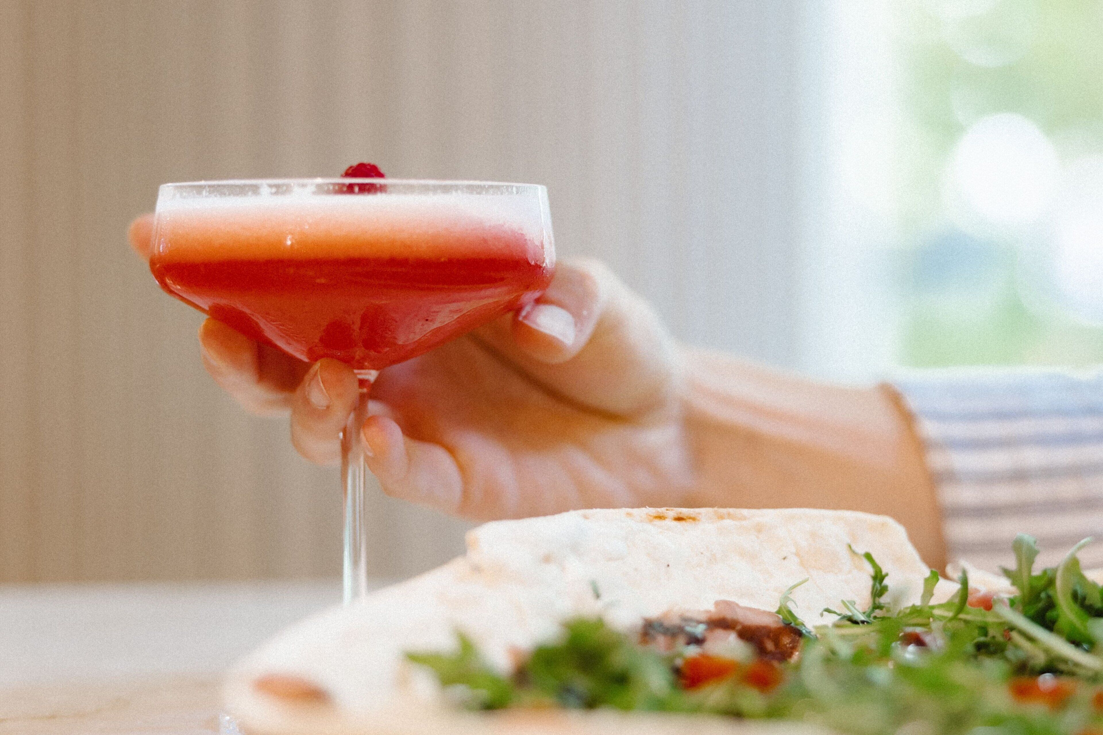 A hand holding a cocktail glass with a red drink, a flatbread with greens and a red sauce, and a blurred background.