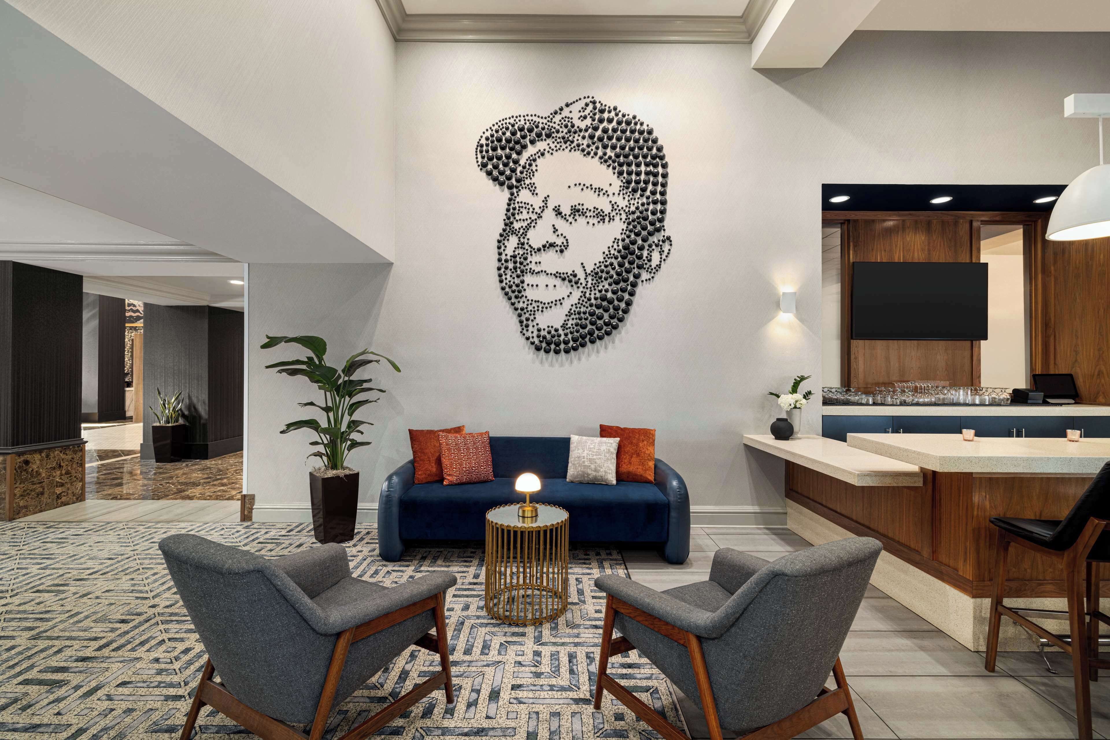 Un moderno vestíbulo de hotel con un sofá azul, dos sillones, una mesita de centro redonda, una gran obra de arte en la pared, una zona de bar y una planta en maceta.