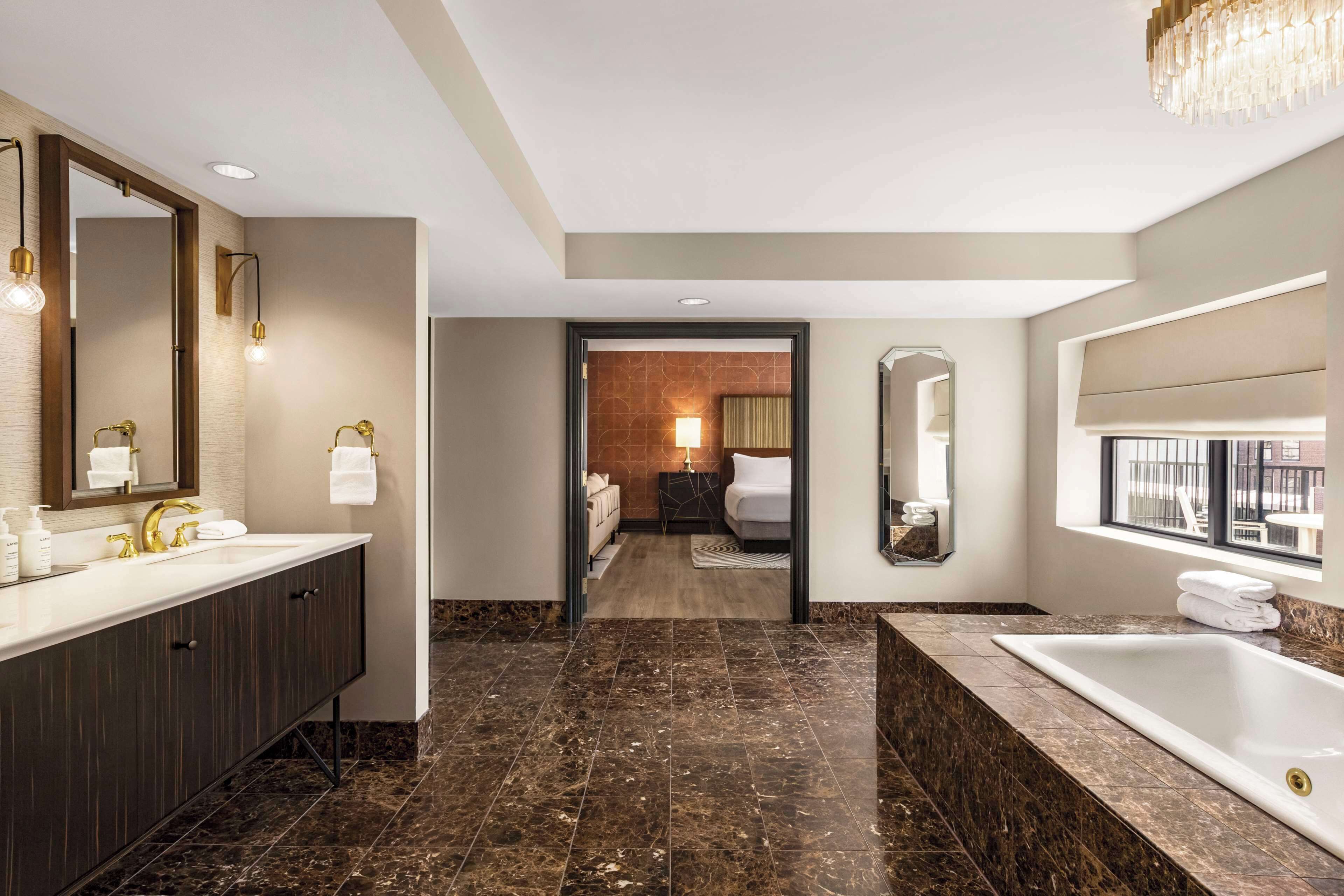 A modern bathroom with a large mirror, dual sinks, a bathtub, and a chandelier.
