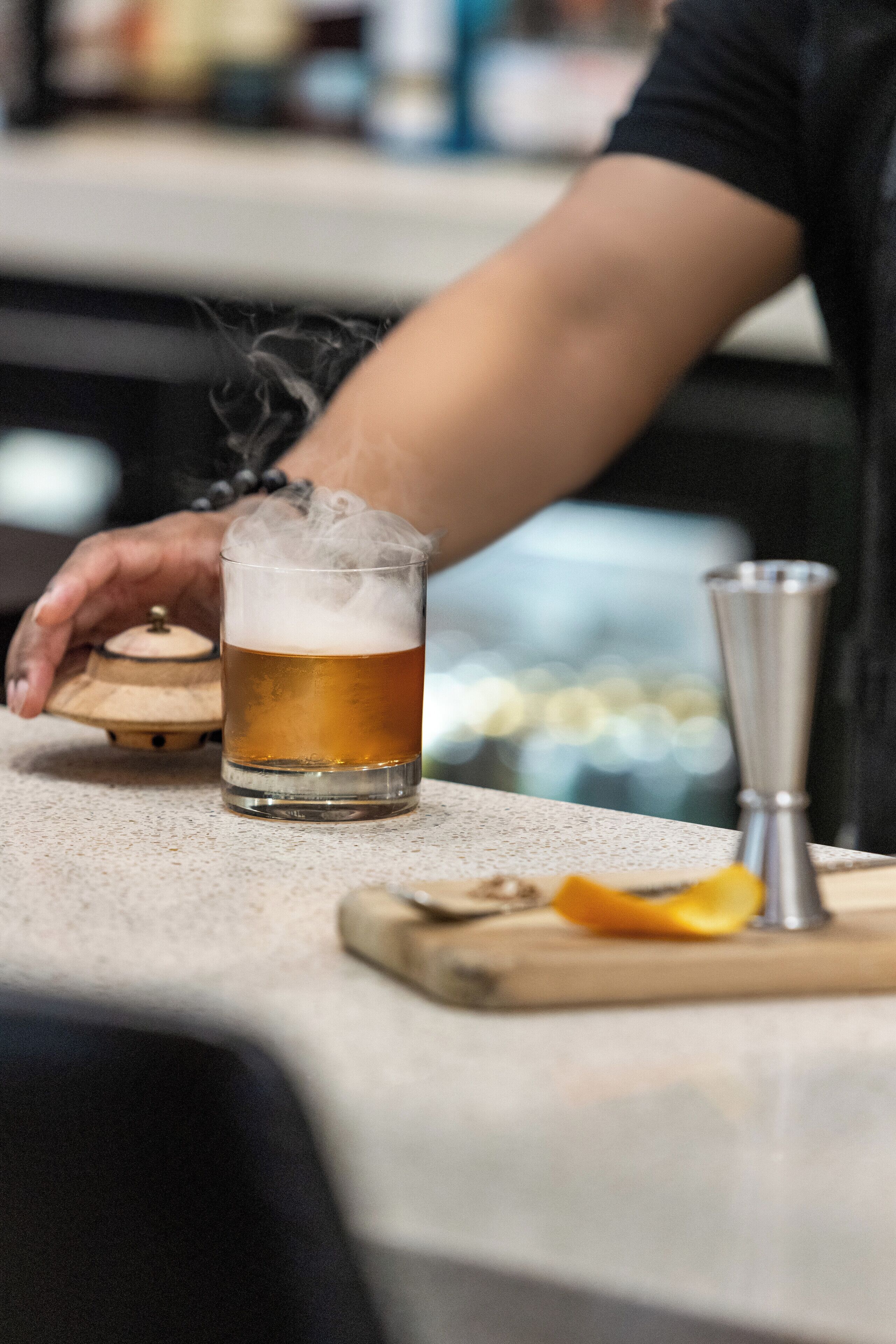 A person pouring a drink into a glass, with a shaker and a wooden container on the counter.