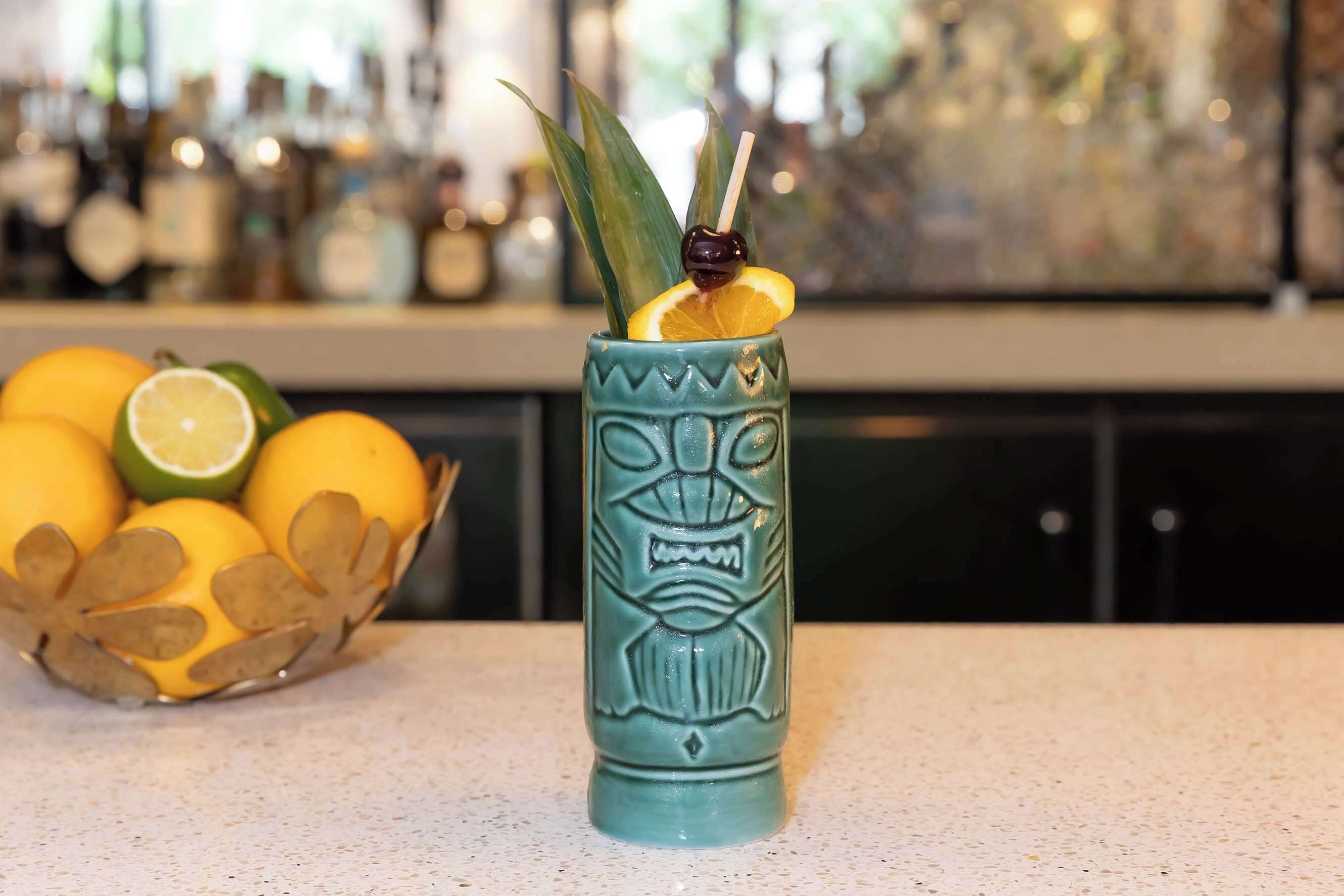 A tiki mug with a carved face, a slice of lime, and a cherry on a stick, placed on a countertop with a bowl of fruit in the background.