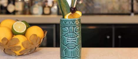 A tiki mug with a carved face, a slice of lime, and a cherry on a stick, placed on a countertop with a bowl of fruit in the background.