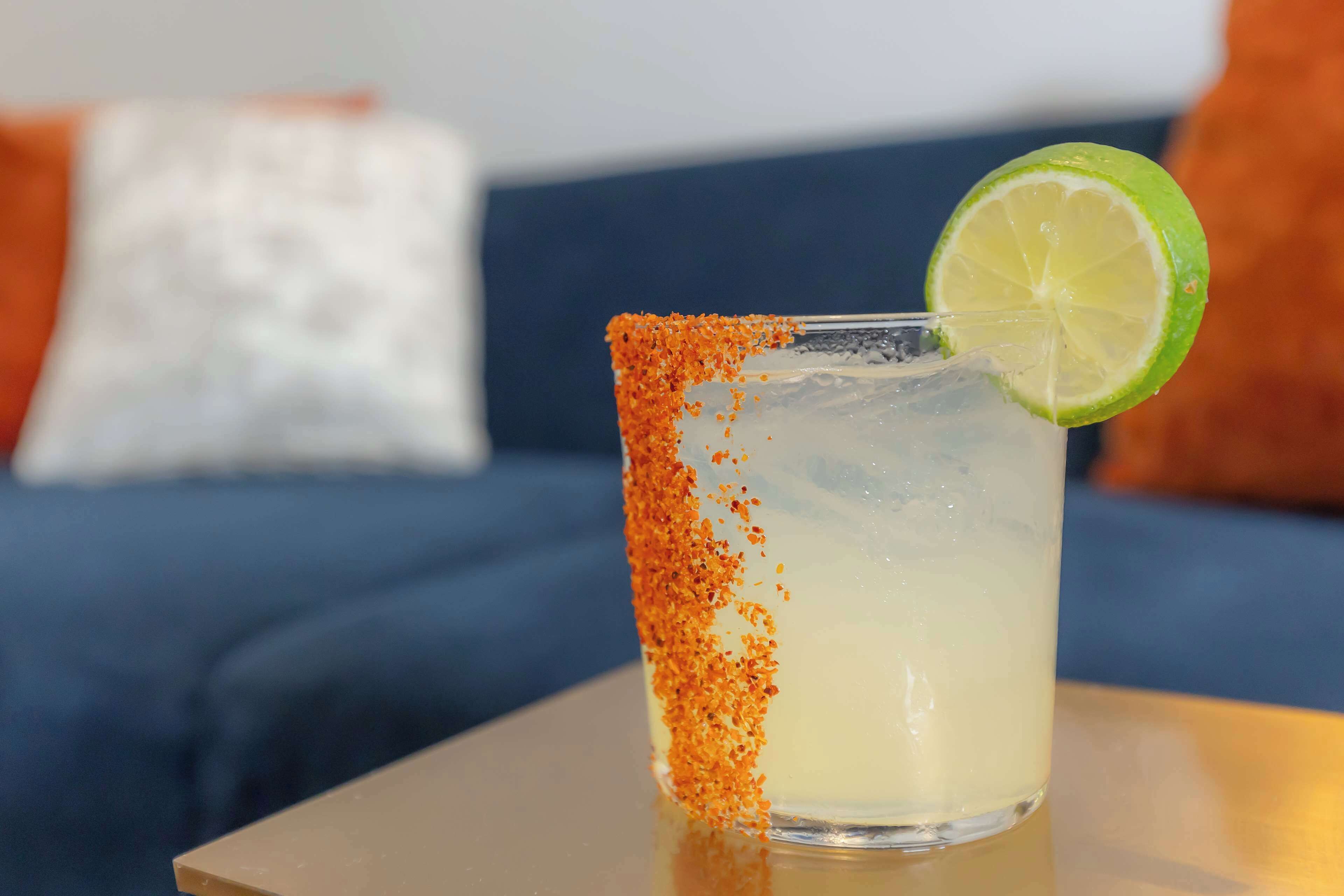 A glass with a salted rim and a lime garnish, placed on a table with a blue couch in the background.
