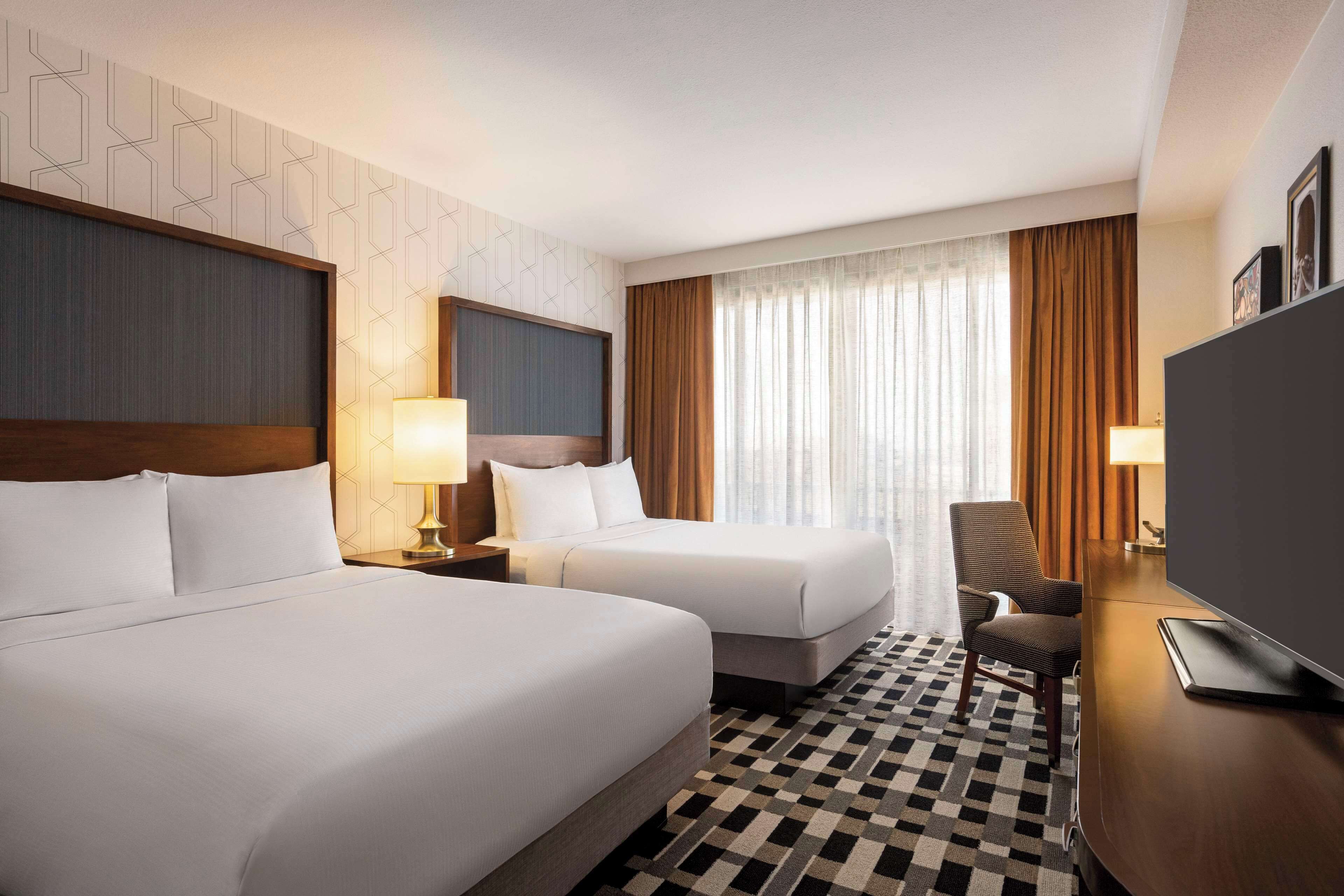 Ein Hotelzimmer mit zwei Betten, einem Schreibtisch, einem Sessel, einem Fernseher und einem gemusterten Teppich.