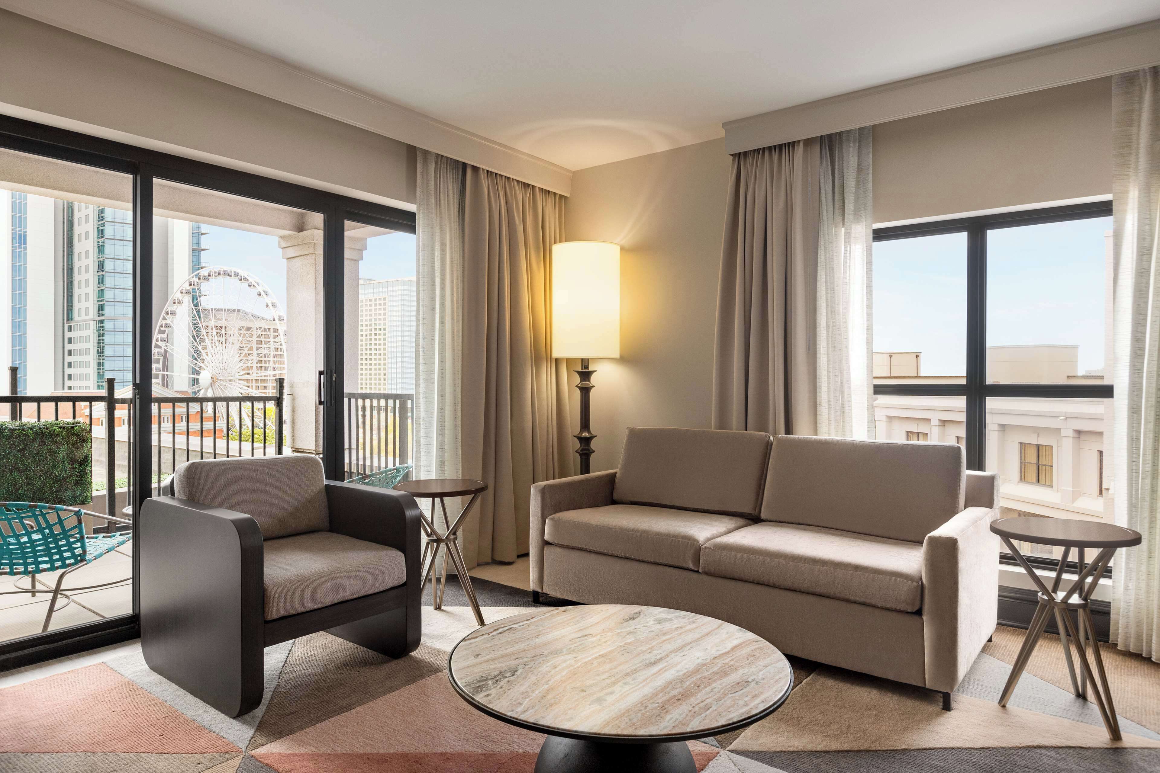 A modern hotel room with a sofa, armchair, and round table. A lamp is on the table, and the room has large windows with a view of a Ferris wheel.
