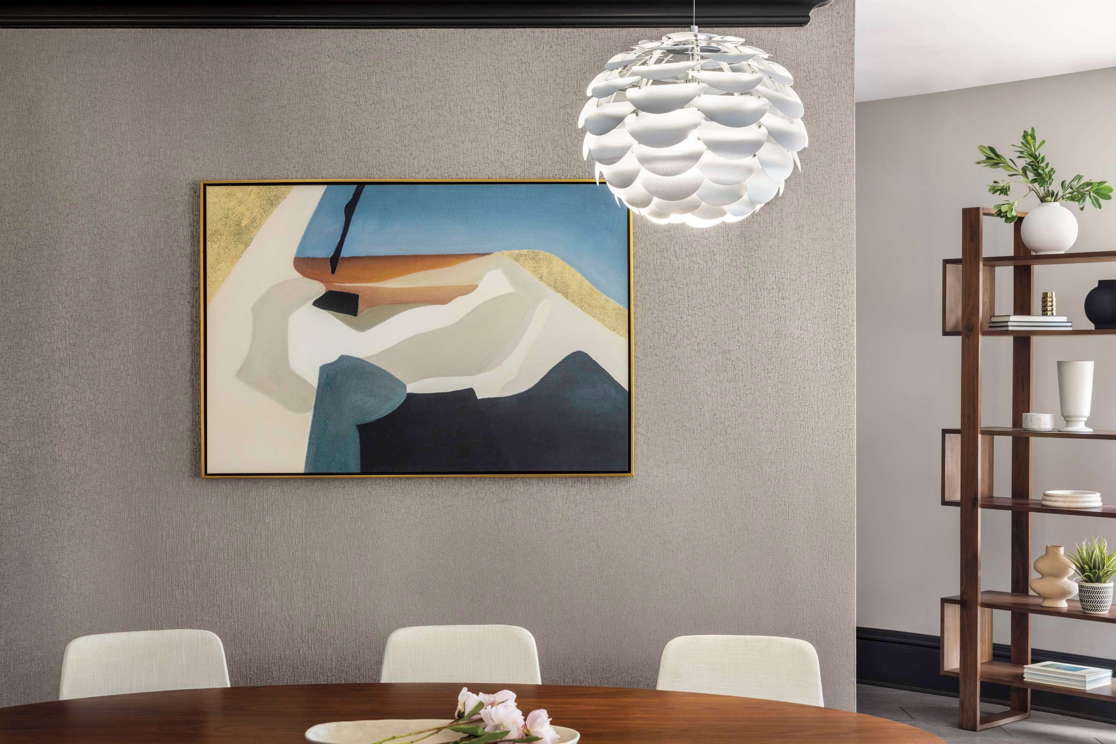 A dining area with a round table, chairs, a painting on the wall, a white pendant light, and a wooden bookshelf with decorative items.