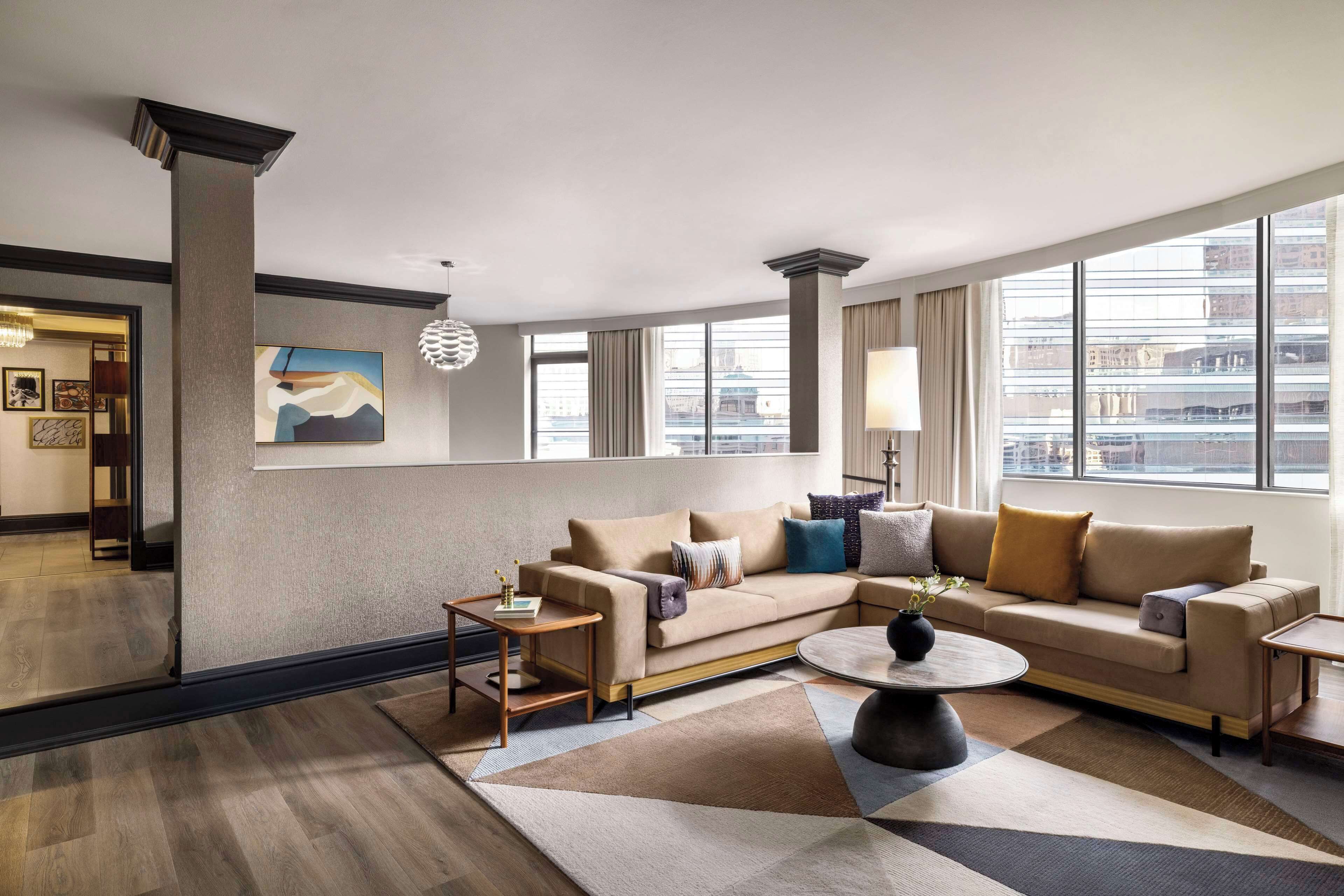A modern hotel room with a large window, a beige sofa, a round coffee table, and a decorative lamp.