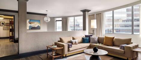 A modern hotel room with a large window, a beige sofa, a round coffee table, and a decorative lamp.
