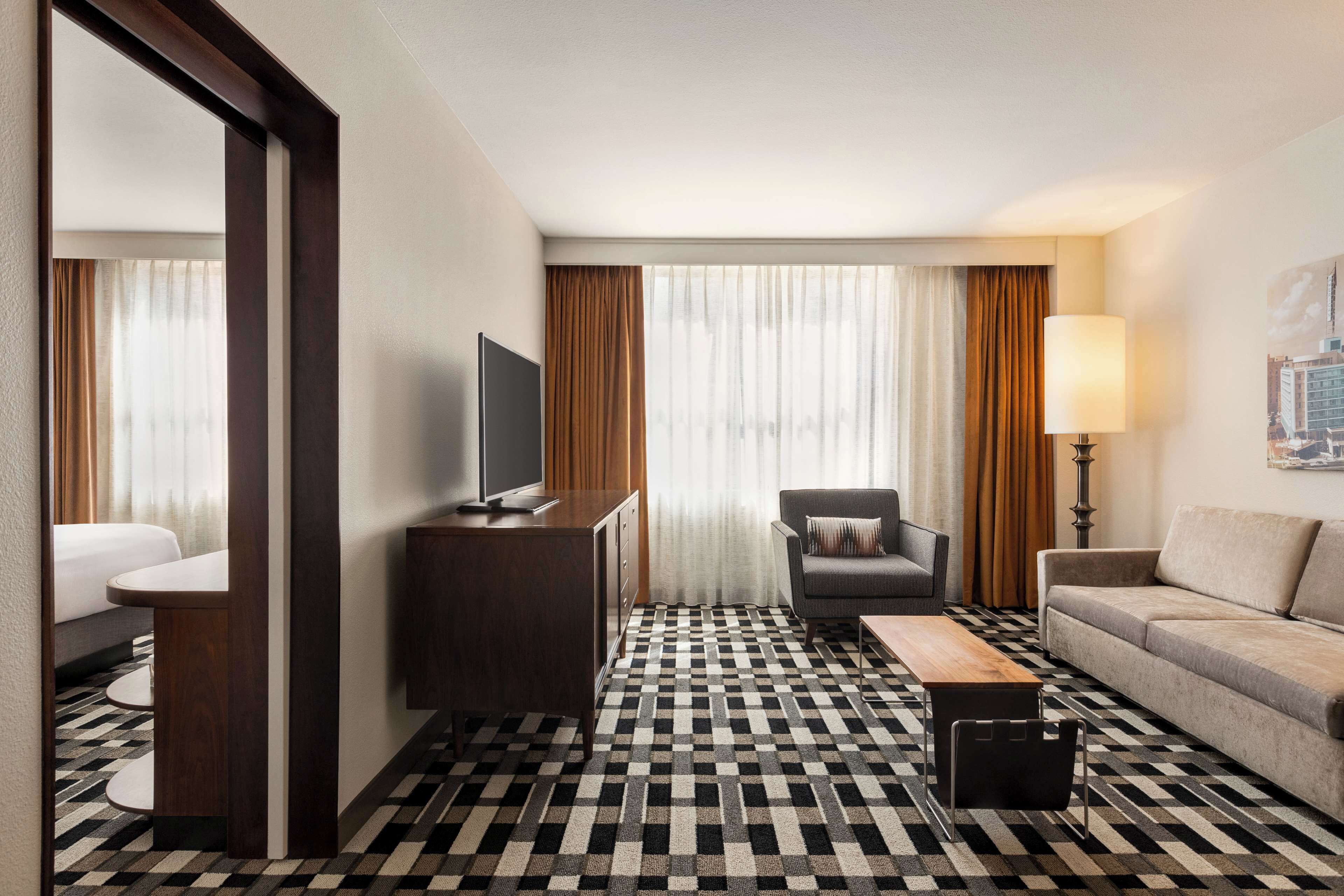 A hotel room with a checkered floor, a sofa, a chair, a TV on a wooden stand, a lamp, and a painting on the wall.