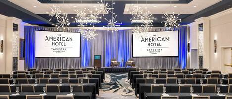 A conference room with rows of chairs and tables, large screens displaying 'The American Hotel Tapestry Collection by Hilton', and a stage with curtains.