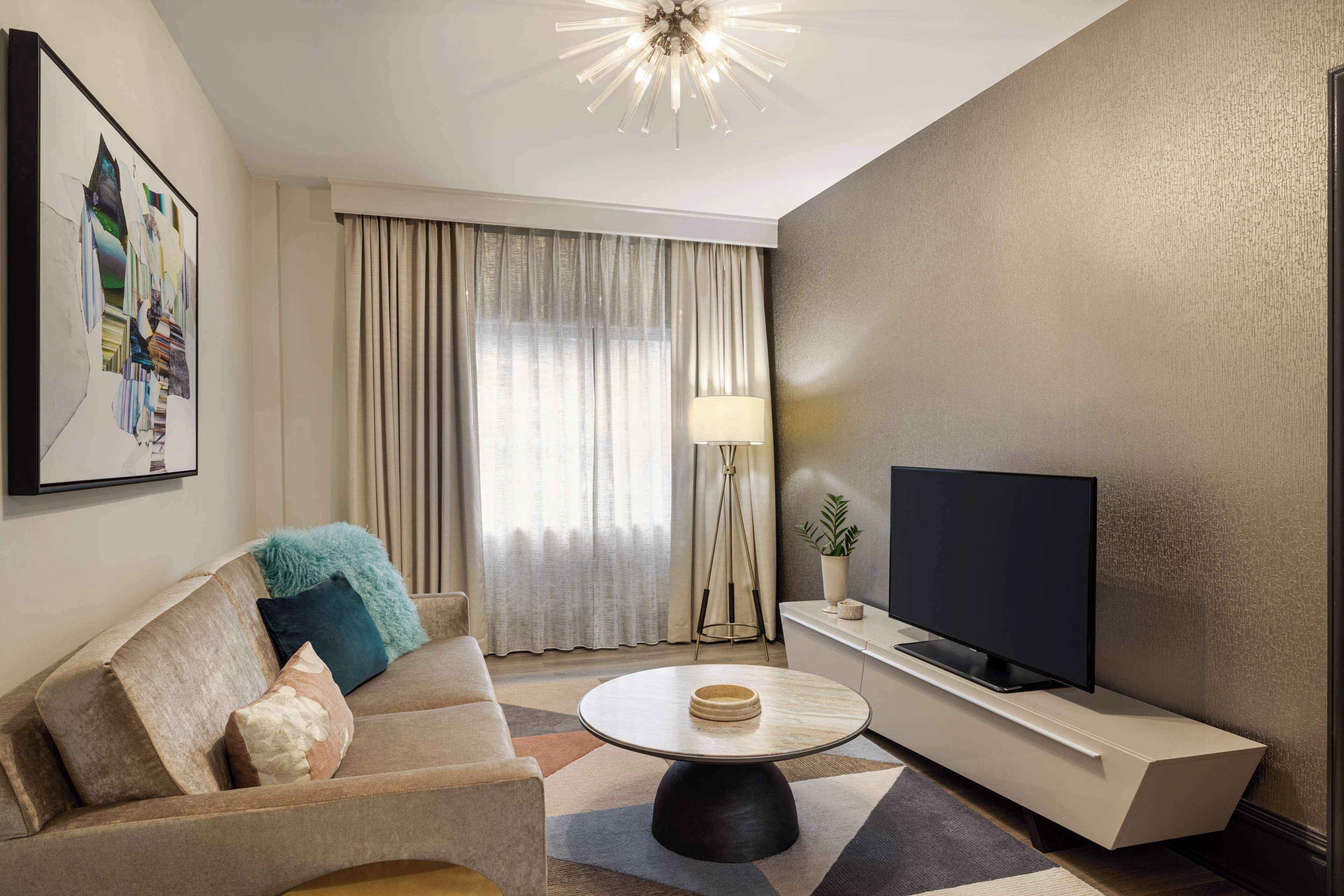 Una sala de estar moderna con un sofá, una mesa redonda de centro, un televisor de pantalla plana sobre un soporte blanco y un ventanal con cortinas.