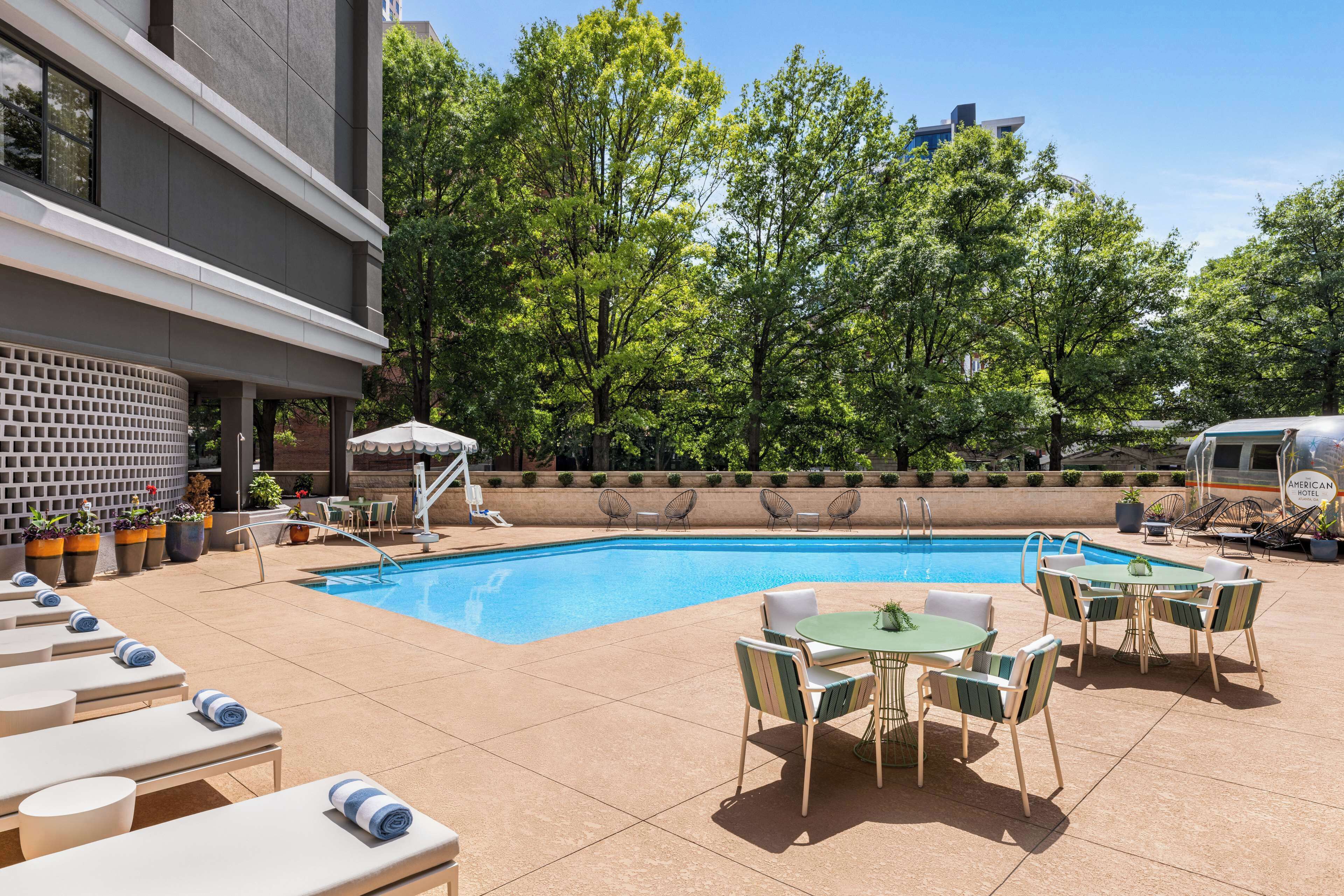 A pool area with lounge chairs, a table, and chairs, surrounded by trees and a building.