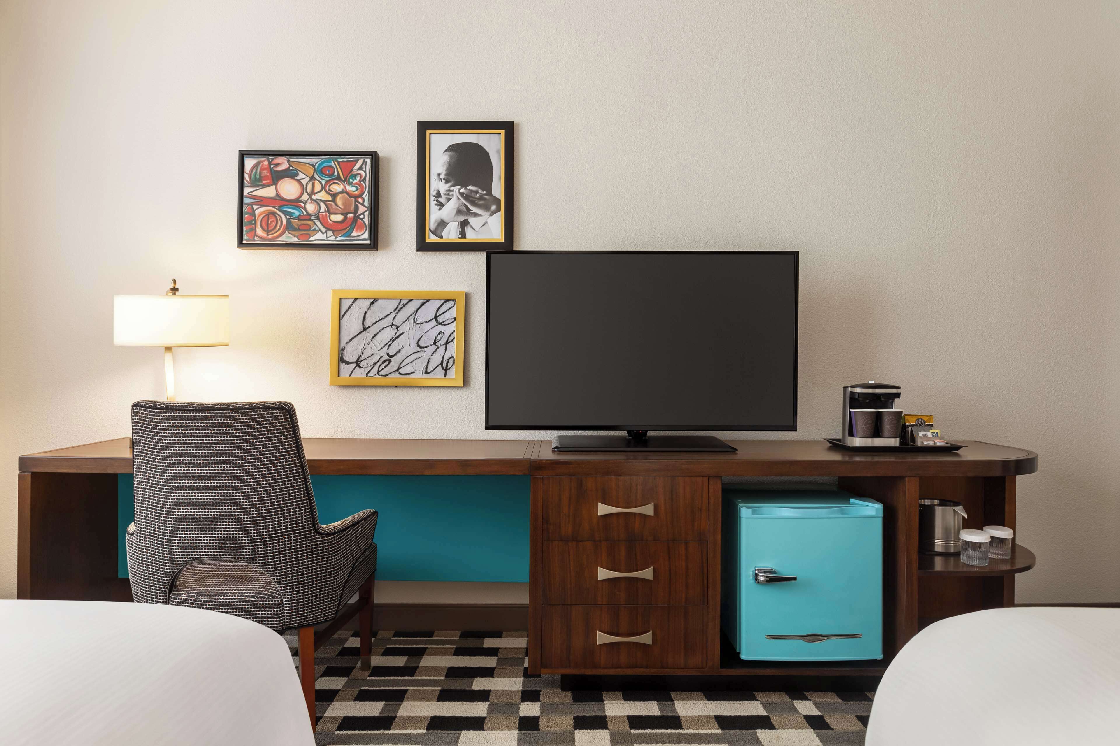 A hotel room with a bed, a desk with a television, a chair, a small refrigerator, and artwork on the wall.