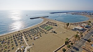 An aerial view of a beach with rows of sunbeds and umbrellas, a pier extending into the sea, and a road alongside the beach.