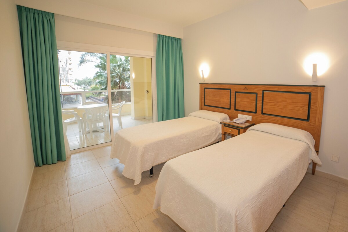 Habitación de hotel con dos camas individuales, cabecero de madera, mesita de noche con teléfono y vistas a un balcón con mesa y sillas.