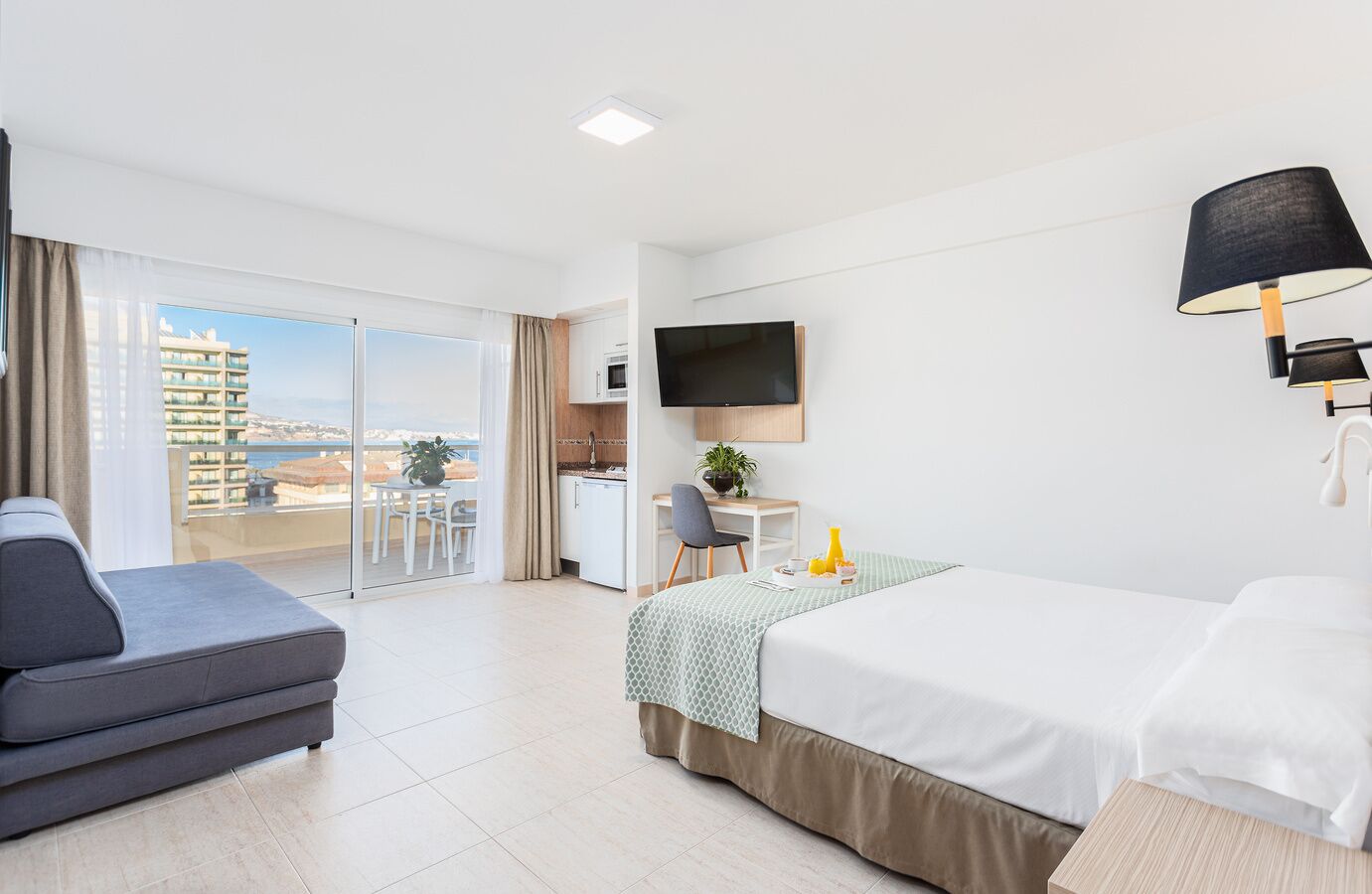 Habitación de hotel con cama, sofá, escritorio con silla, televisor y balcón con vistas a edificios y al mar.