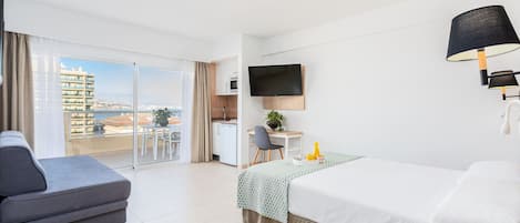 Habitación de hotel con cama, sofá, escritorio con silla, televisor y balcón con vistas a edificios y al mar.