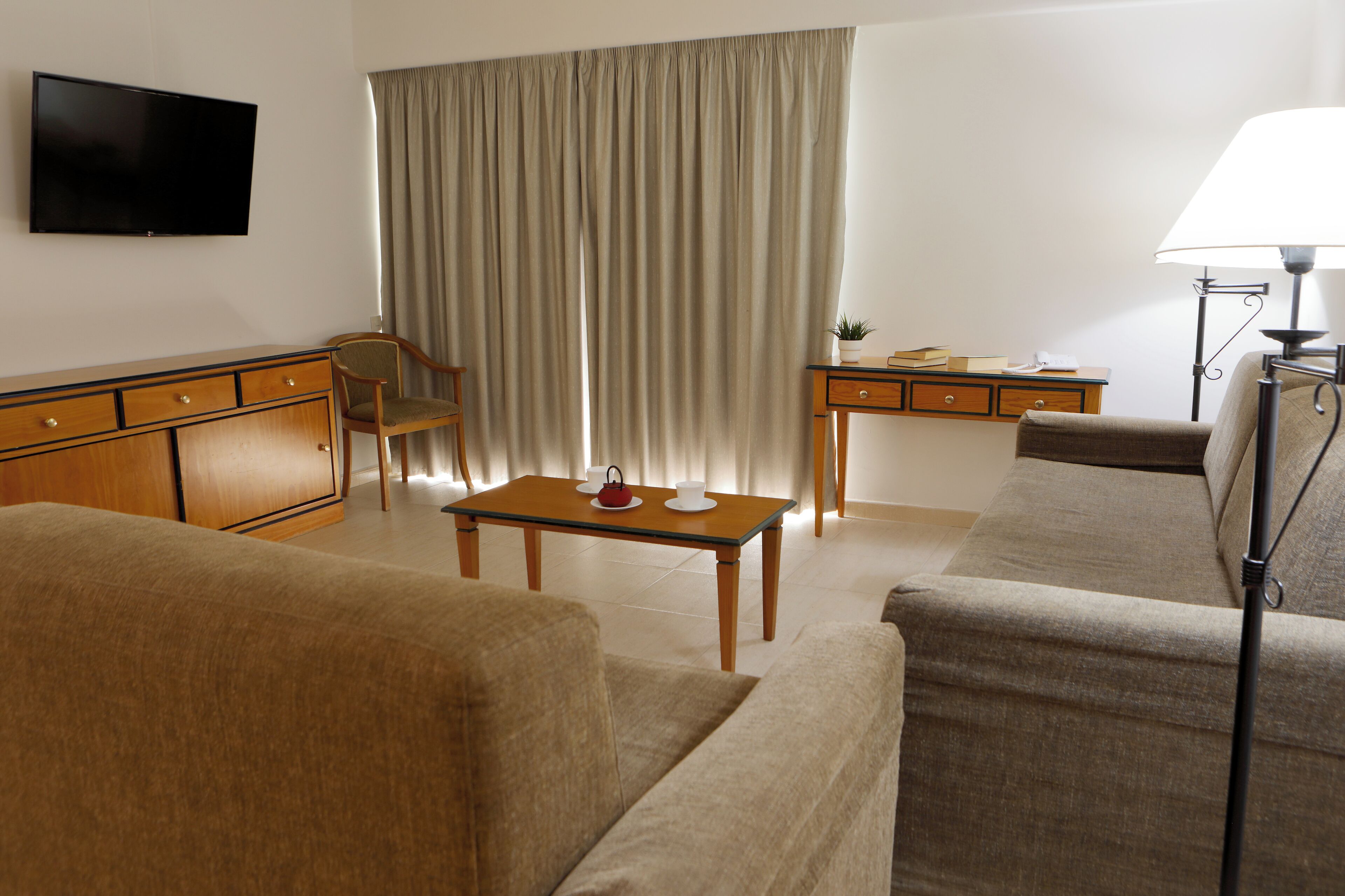 A hotel room with a sofa, a wooden desk, a television, a lamp, and a curtain.