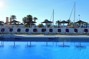 Un espace aménagé au bord de la piscine, avec des chaises longues et des parasols, un ciel dégagé et des palmiers.