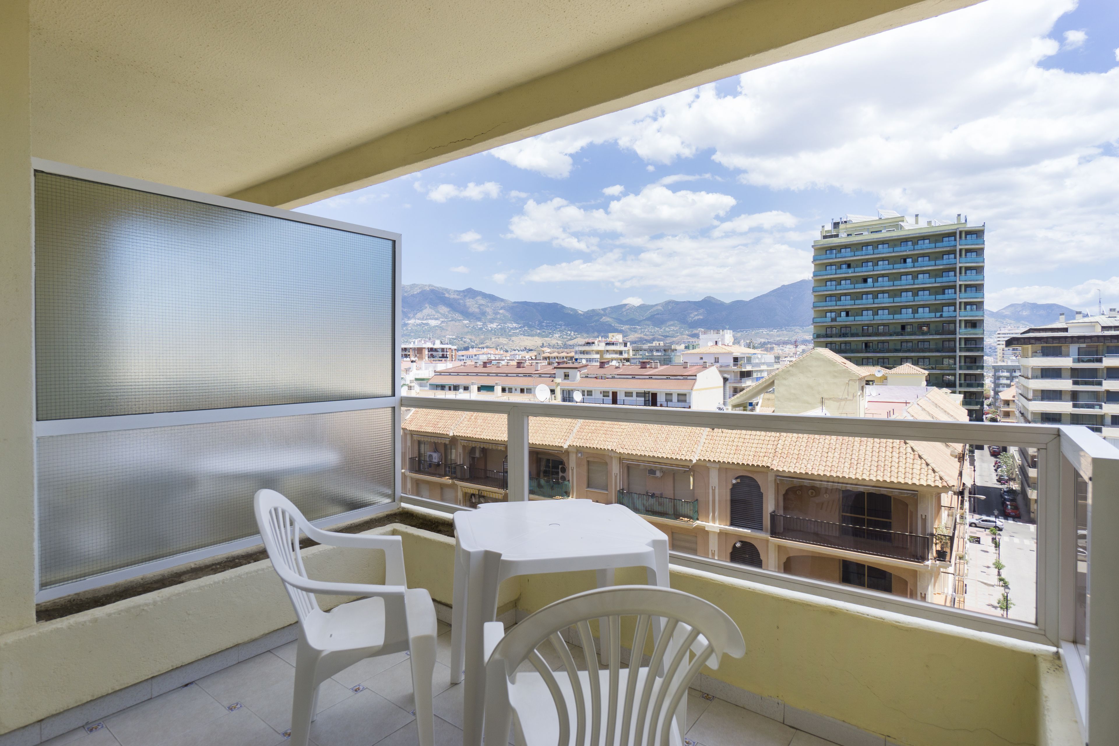 A balcony with a white table and chairs, overlooking a cityscape with buildings and mountains.