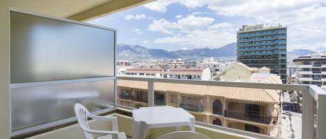 A balcony with a white table and chairs, overlooking a cityscape with buildings and mountains.