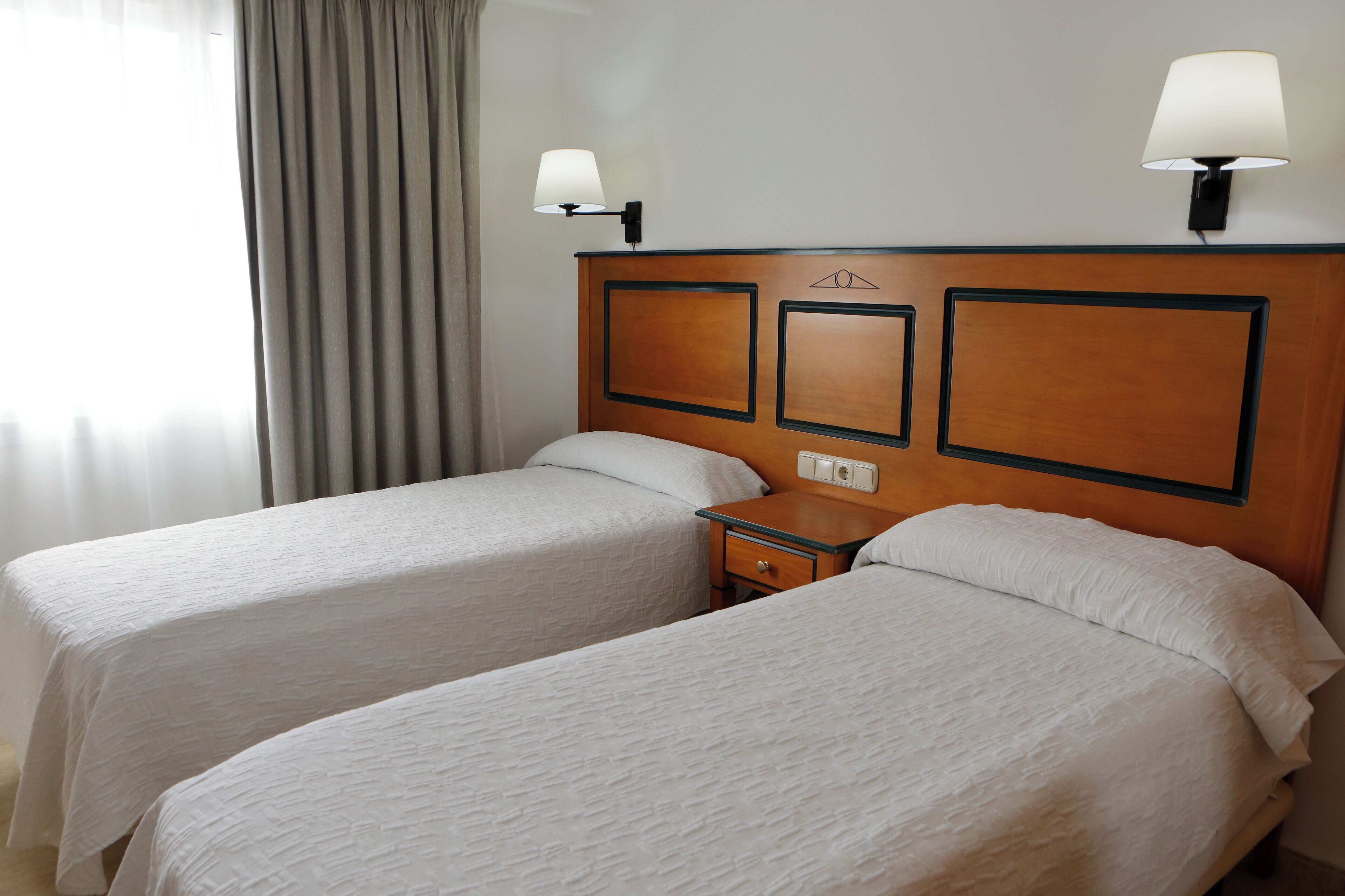 Hotellihuone, jossa on kaksi erillistä sänkyä, yöpöytä, lamppu ja ikkuna, jossa on verhot.