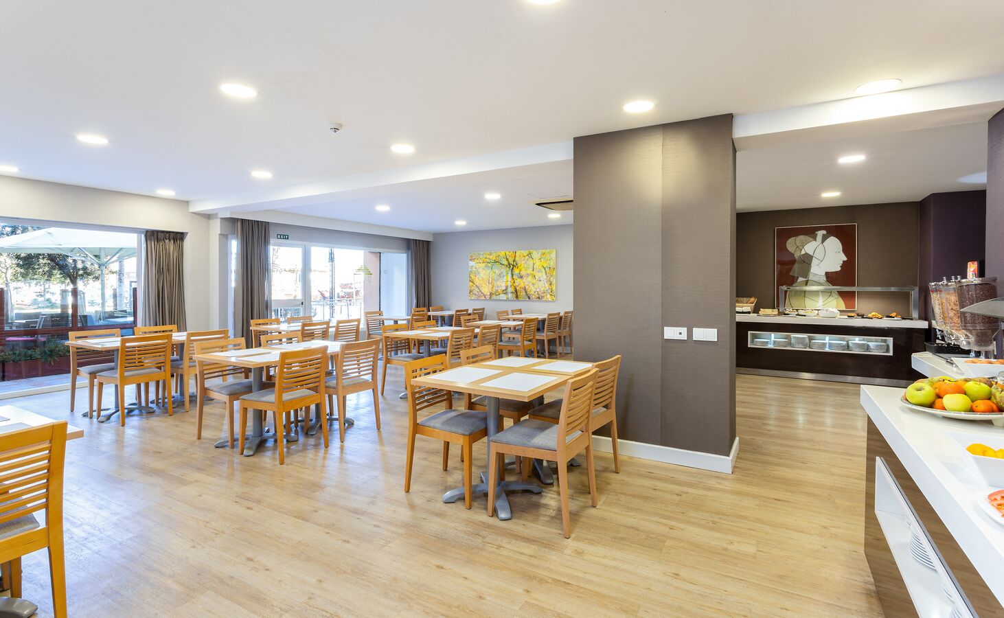 Et moderne spiseområde med træborde og stole, et buffetbord med frugt og et stort vindue med gardiner.