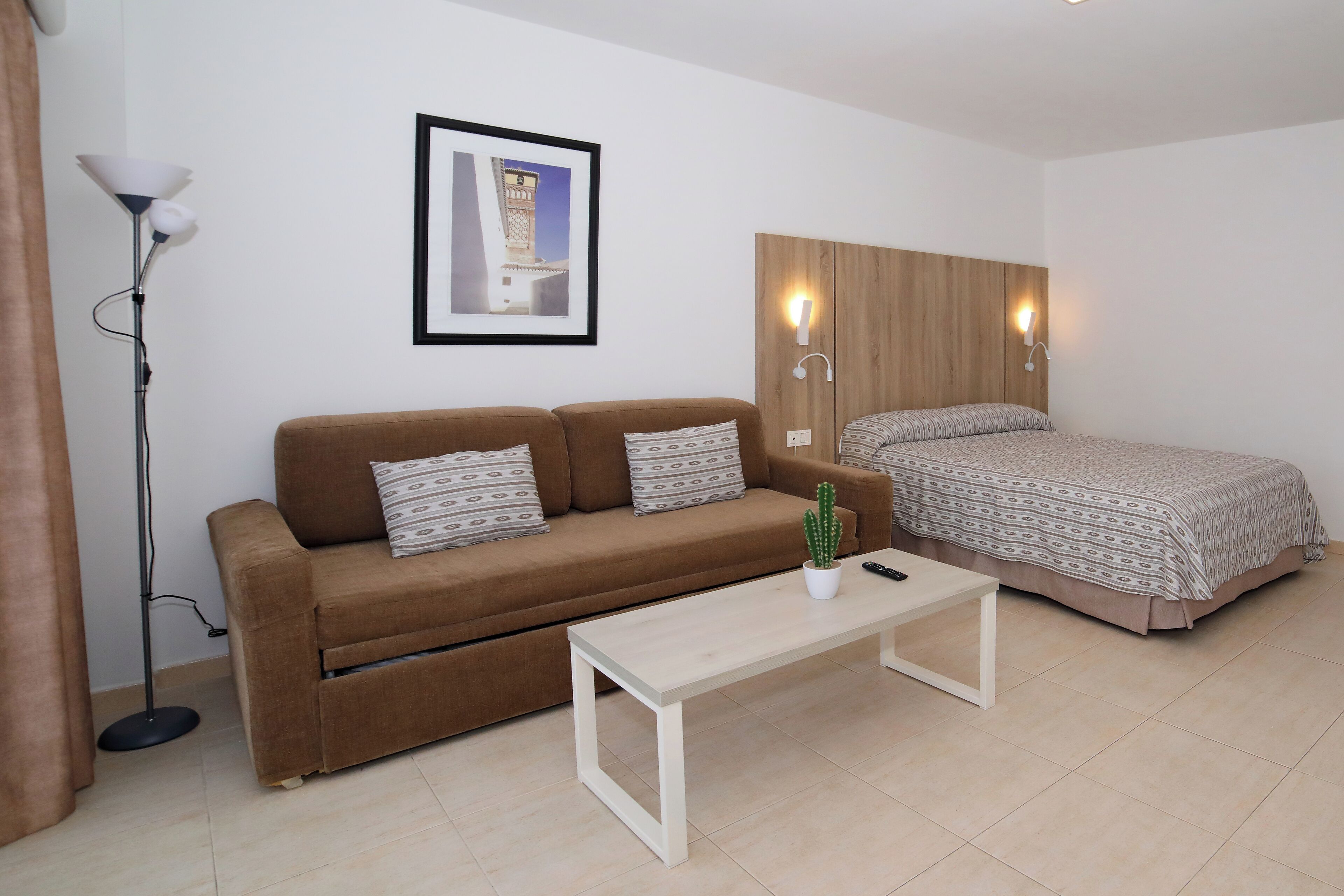 A room with a brown sofa, a white coffee table, a bed with patterned bedding, a framed picture on the wall, and a floor lamp.