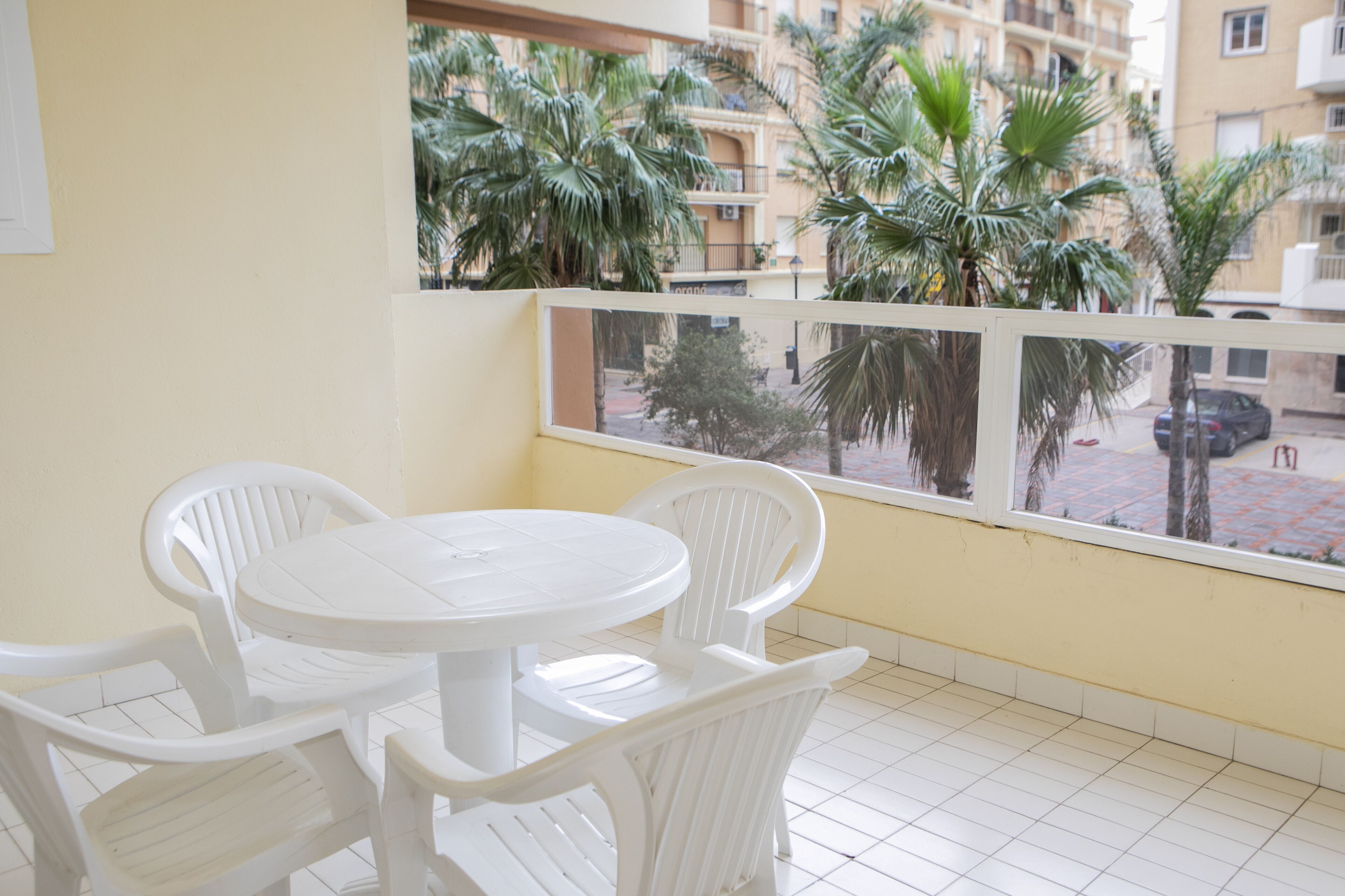 A balcony with white plastic chairs and a table, overlooking a street with palm trees and buildings.