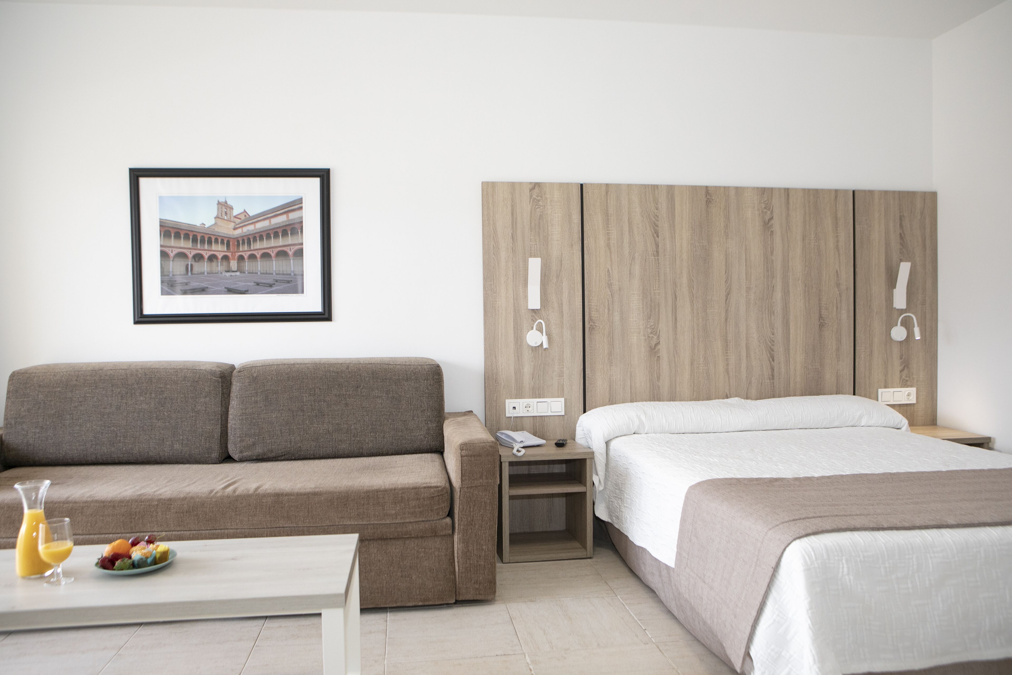 Une chambre d’hôtel avec un canapé, un lit, une petite table sur laquelle se trouvent un verre de jus d’orange et des fruits, et un tableau encadré accroché au mur.
